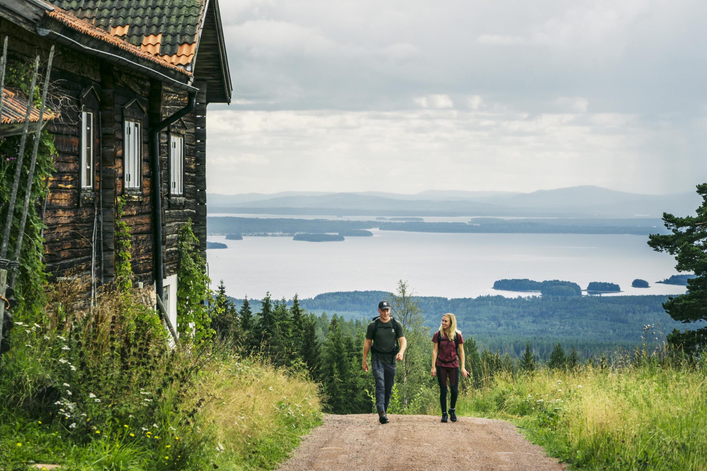 A couple are hiking on a hill next to a wooden house with a view of a lake in the background.