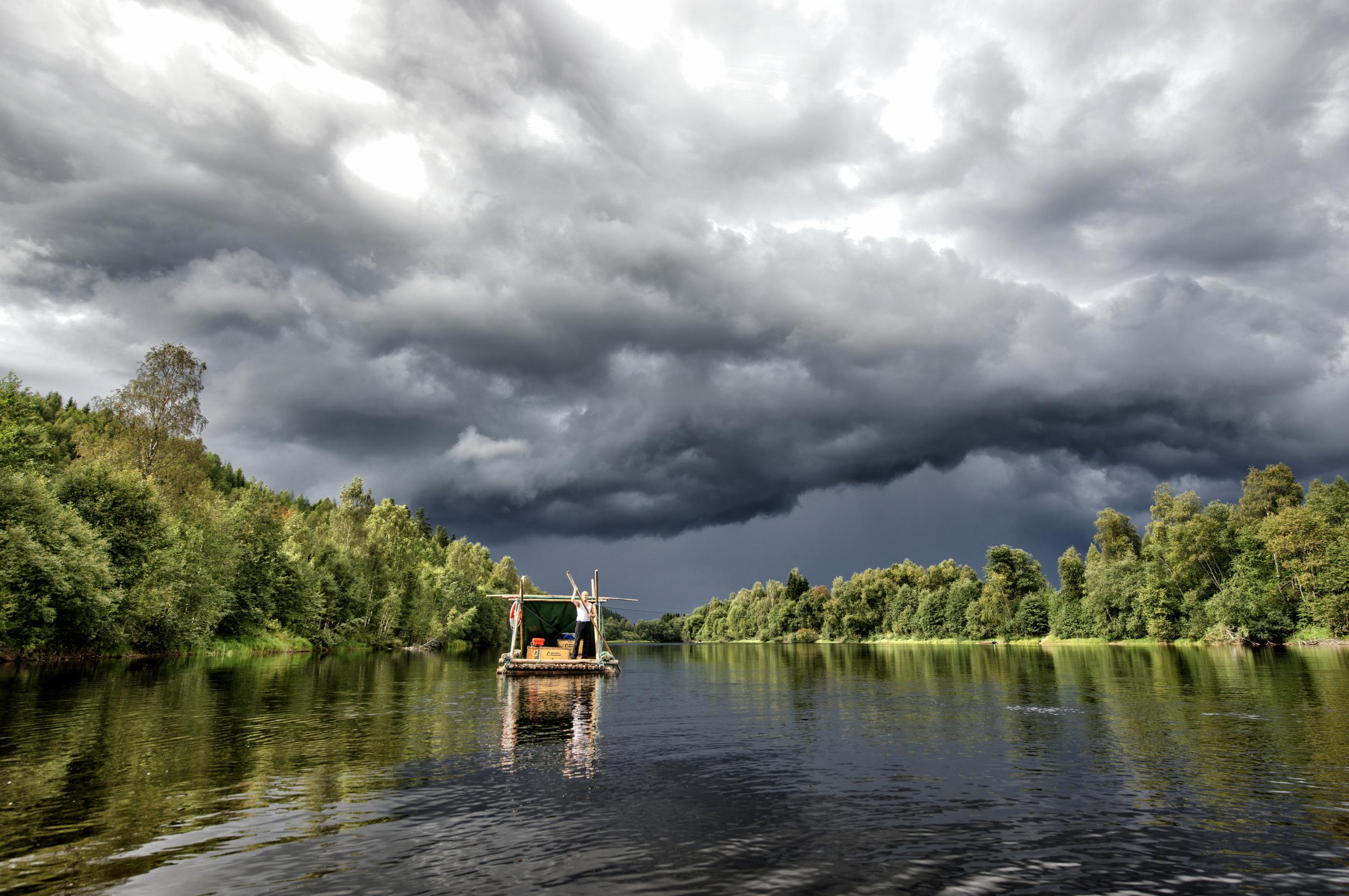 Een houten vlot op een rustige rivier. Op de oever is er veel beplanting. Er hangen donkere wolken boven de rivier.