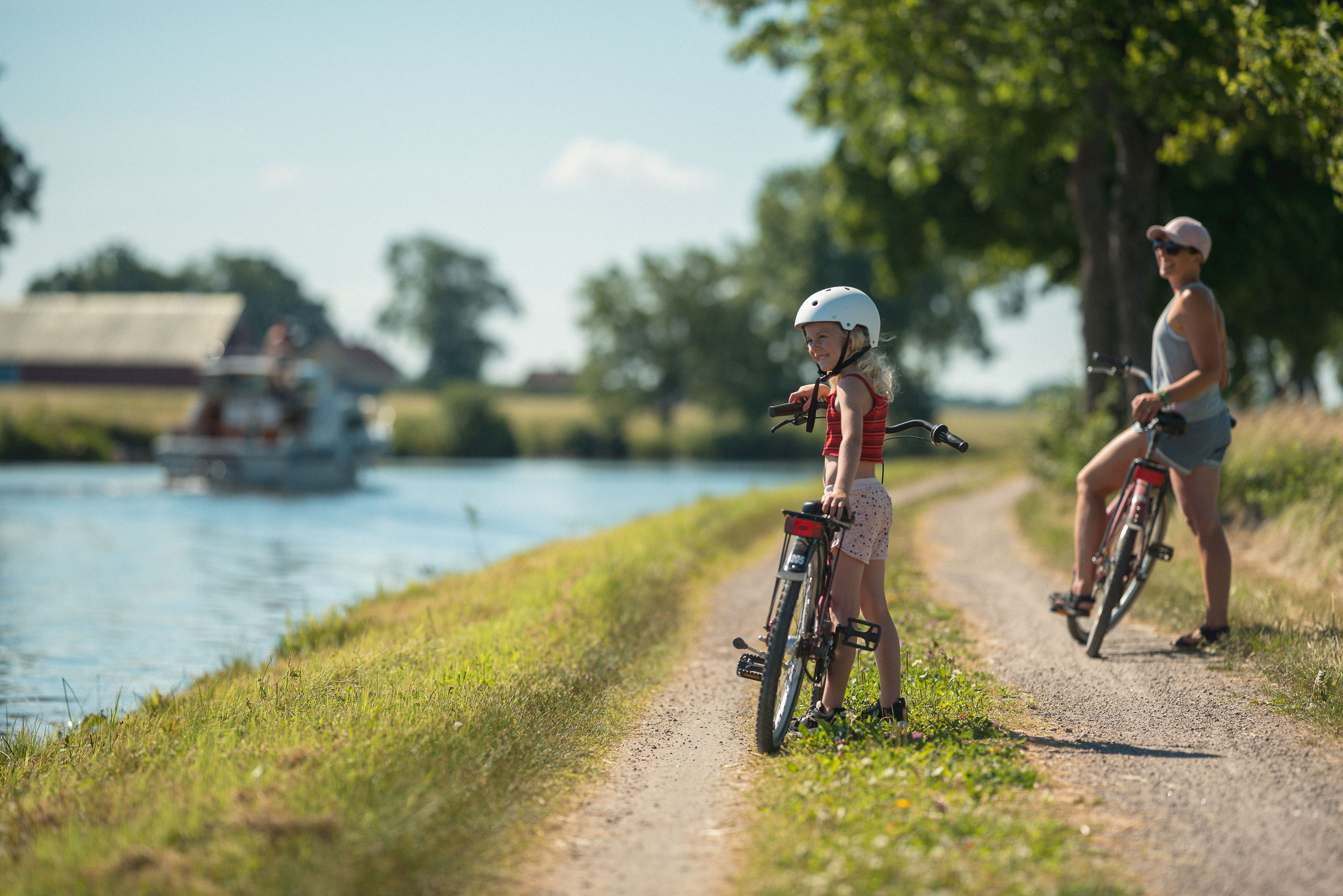 A young girl and an adult woman with bicycles on a gravel path beside the Göta Canal, with a boat cruising in the background on a sunny summer day.