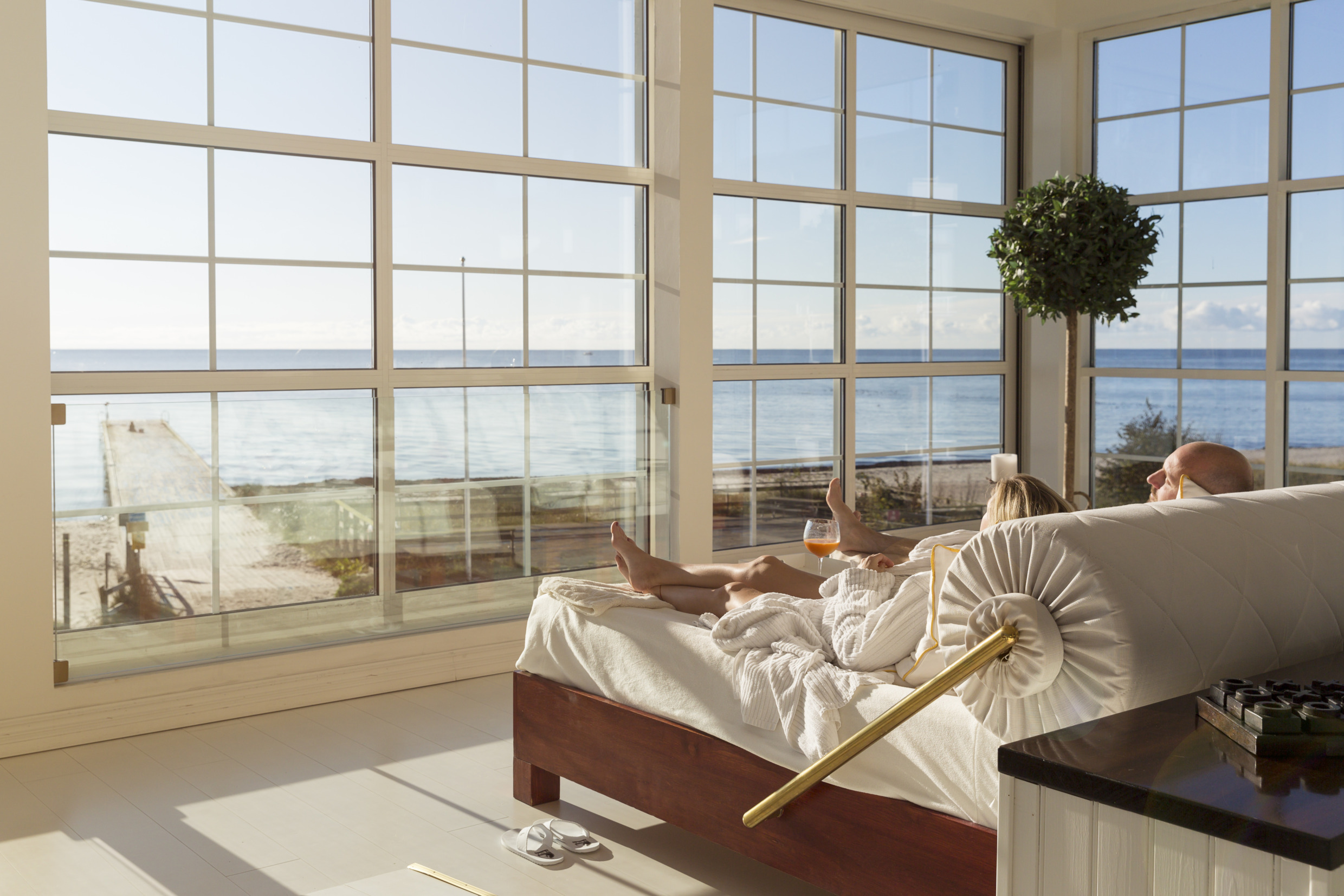 Een stel ligt in een groot bed naast grote ramen met uitzicht op het strand en de zee