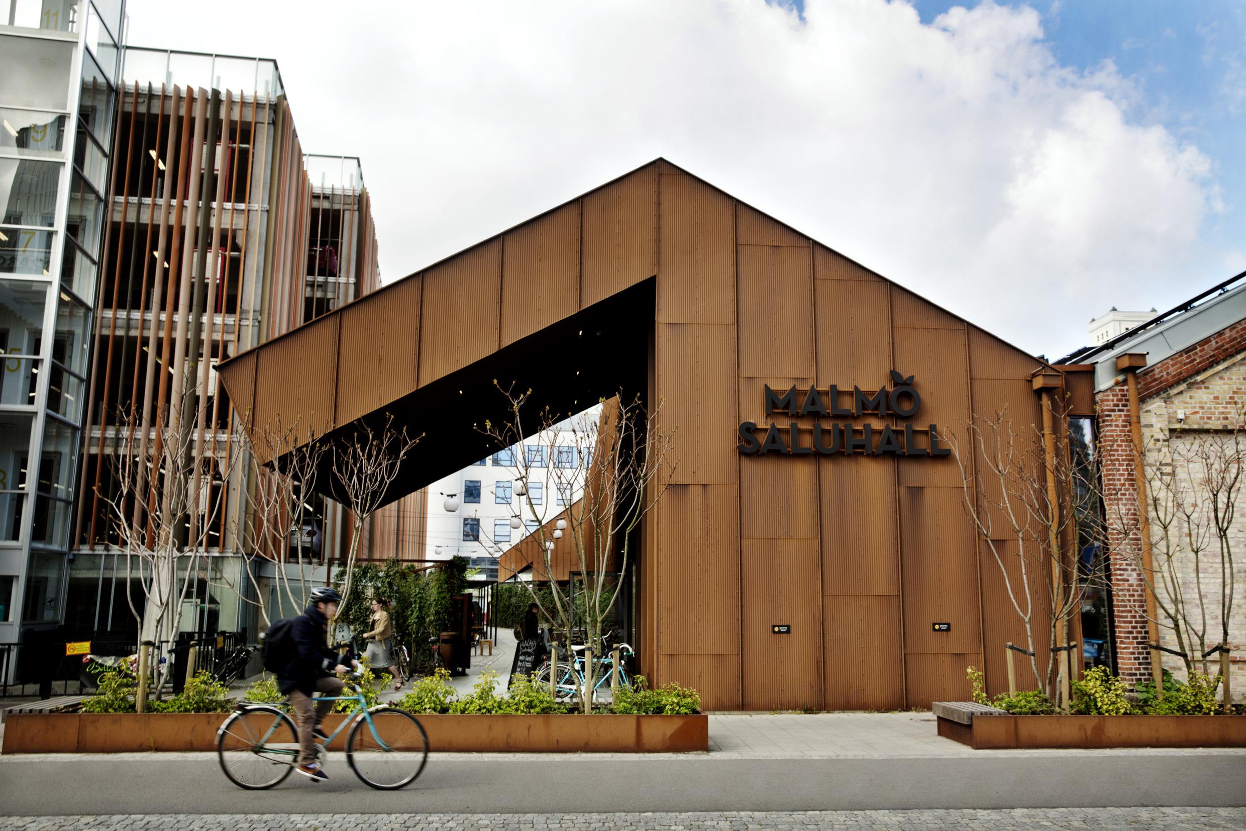Außenansicht der Malmö Saluhall. An der industriell-schicken Markthalle fährt ein Fahrradfahrer vorbei.