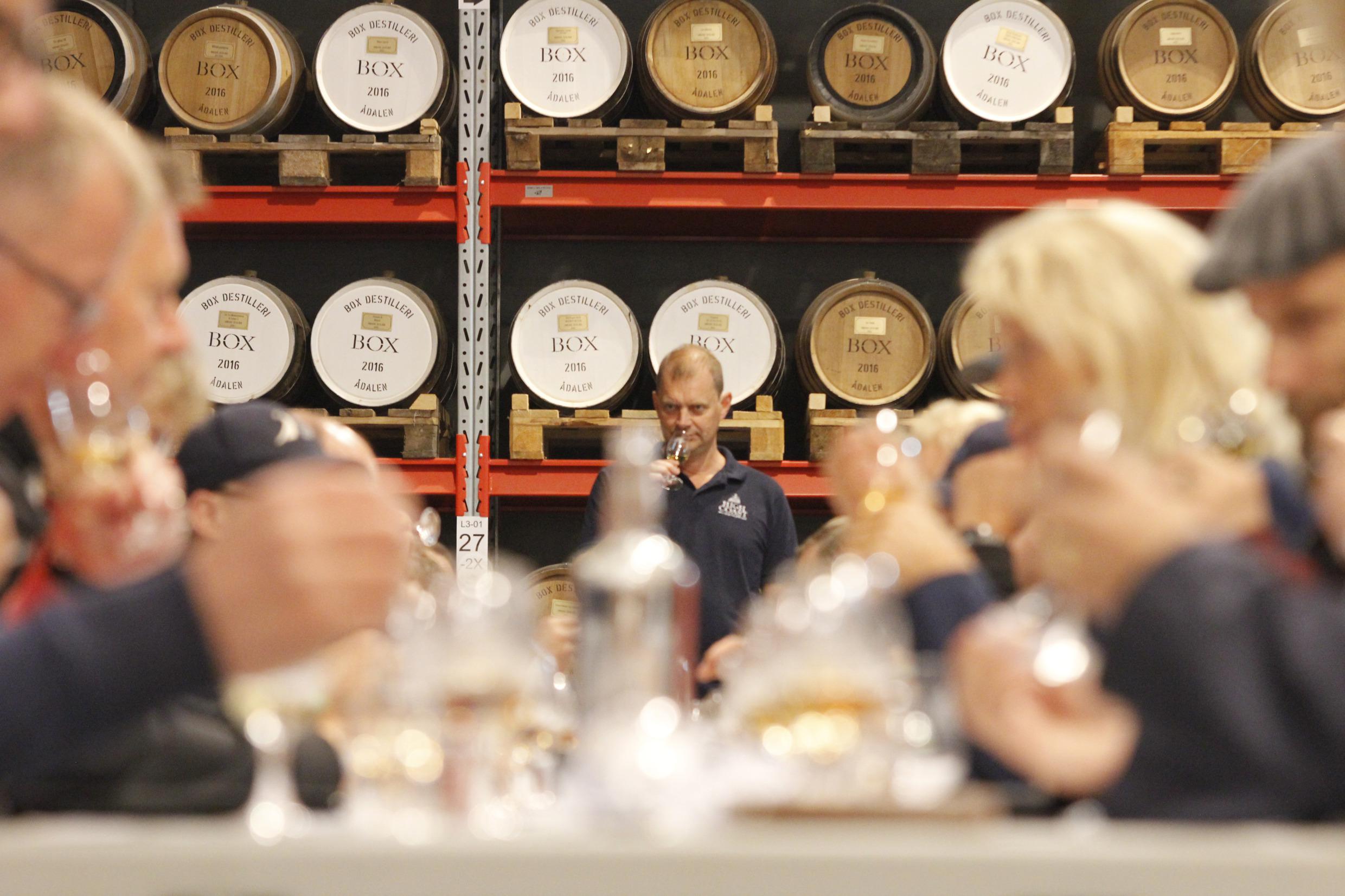 Ein Mann steht am Ende eines langen Tisches und riecht an seinem Whiskey. Eine Gruppe von Menschen, die bei einem Festival am Tisch sitzen und Whisky probieren.