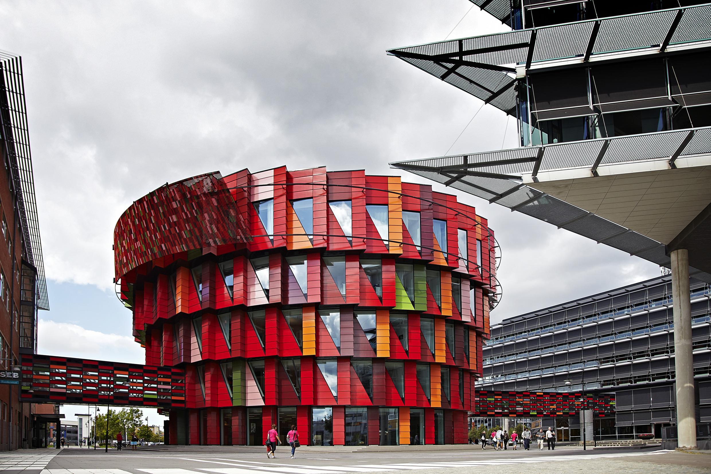 Een kleurrijk, veelal rood, rond gebouw met driehoekige ramen. Er lopen wat mensen naast het gebouw.