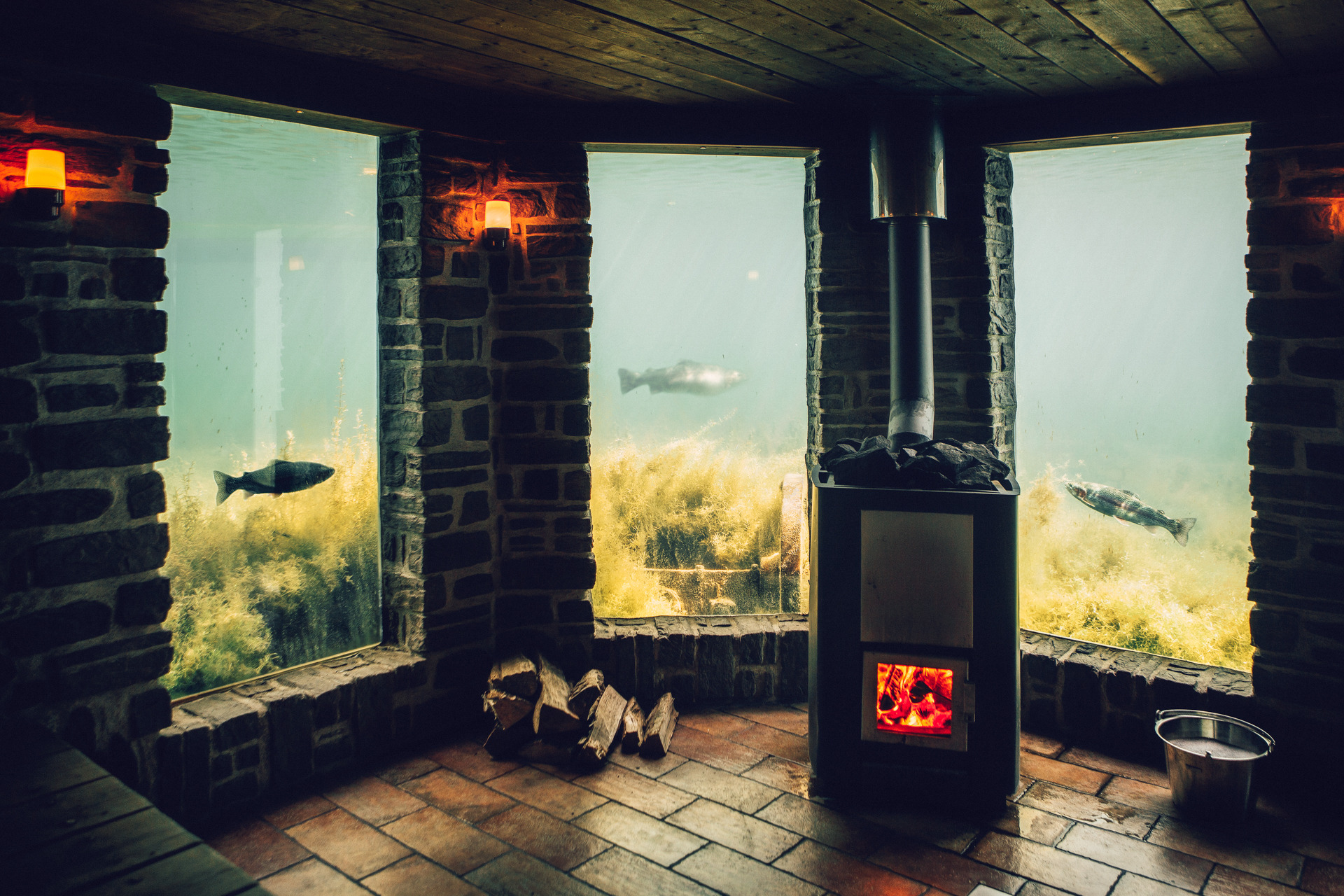 Interieur van een onderwatersauna in Ästad Vingård, met een houtkachel, stenen muren en grote ramen die een onderwaterzicht bieden, met vissen die tussen de waterplanten zwemmen.