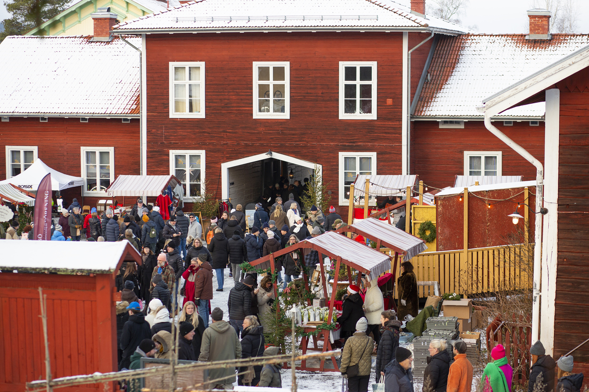 Crowds browsing decorated market stalls in the snow-covered courtyard of red wooden buildings at Stenegård’s Christmas market.