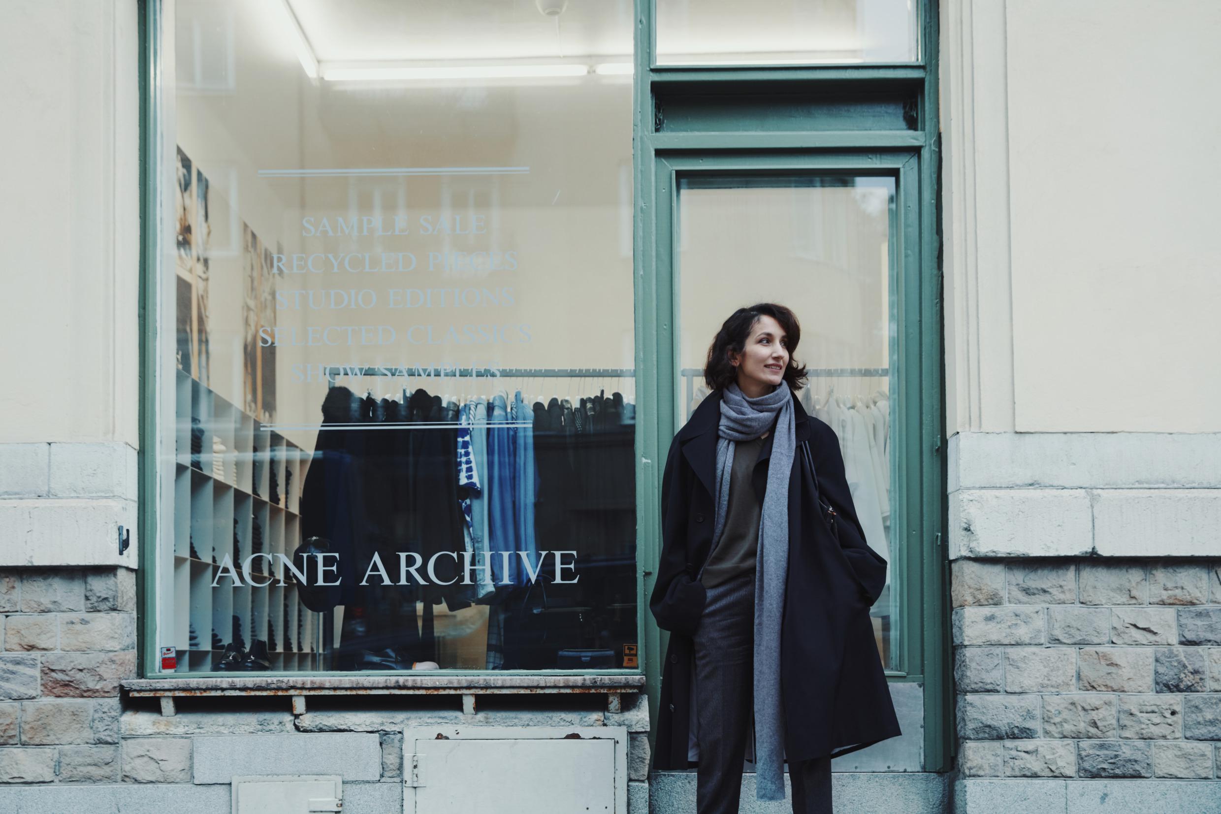 A woman is standing on the street outside an Acne store in Stockholm. Racks of clothes can be seen through the window.