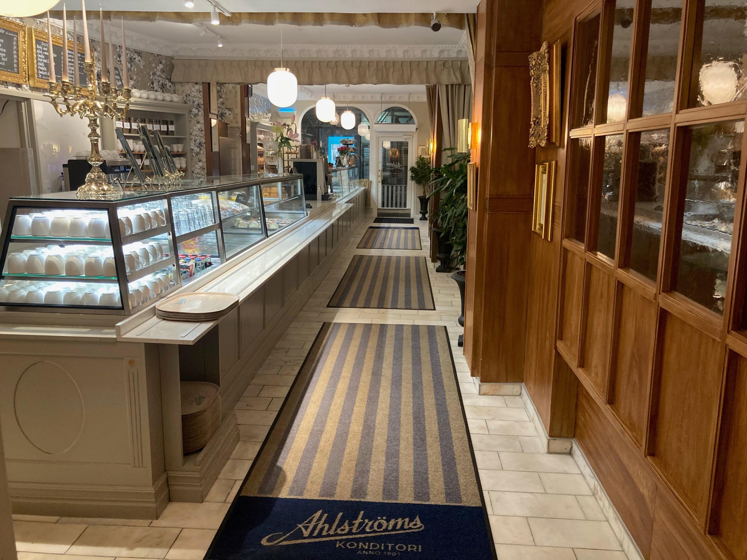 In een leeg café. Een toonbank met glazen vitrines, een kandelaar die op de toonbank staat, een wand in hout en glas aan de zijkant en gestreepte tapijten op de vloer.