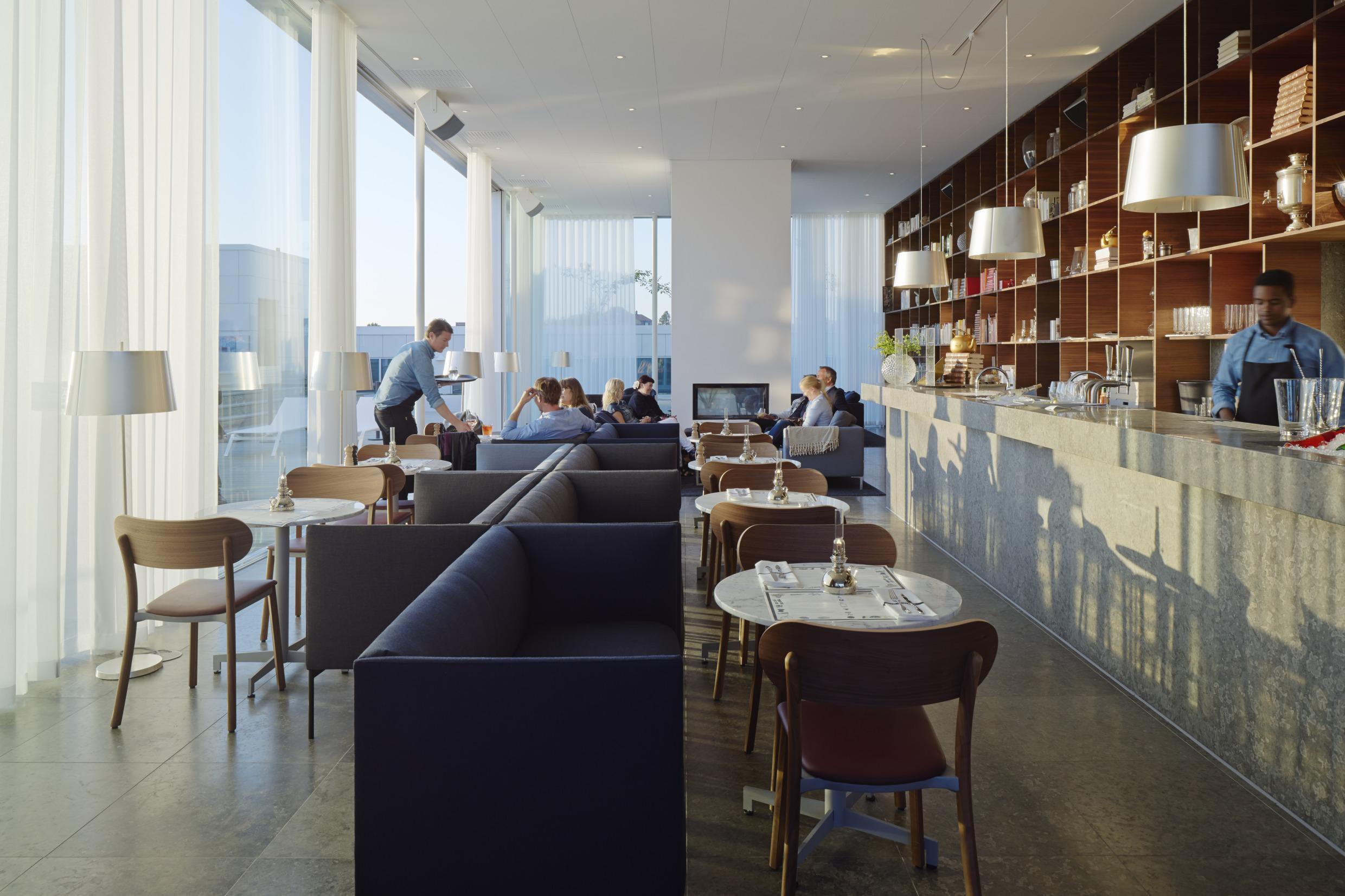 The roof bar at PM & Vänner. Behind a long bar you see a bartender working. There are sofas, chairs and tables and there some guests sitting in the far end.