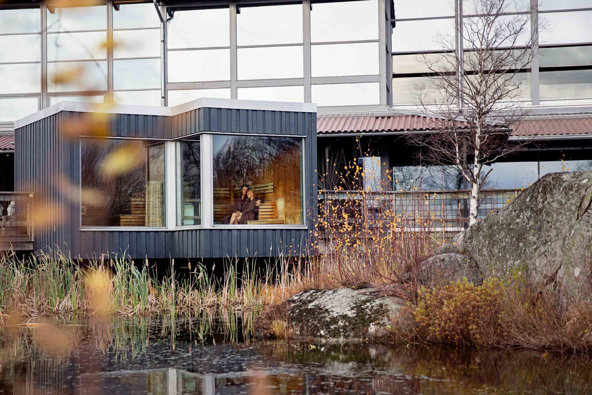 Een houten huis met grote panoramische ramen tegenover een klein meer.