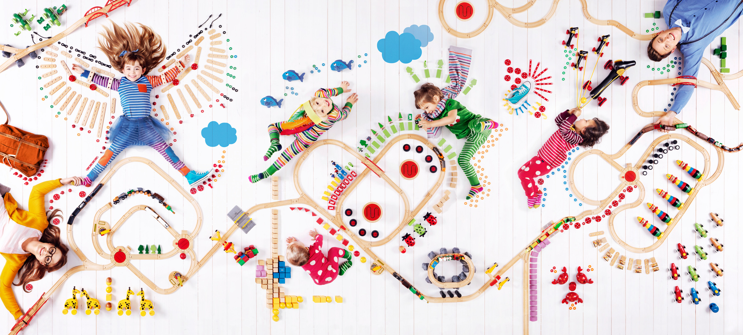 Overhead view of children and adults playing with colourful BRIO train sets arranged in imaginative patterns on a white wooden floor.