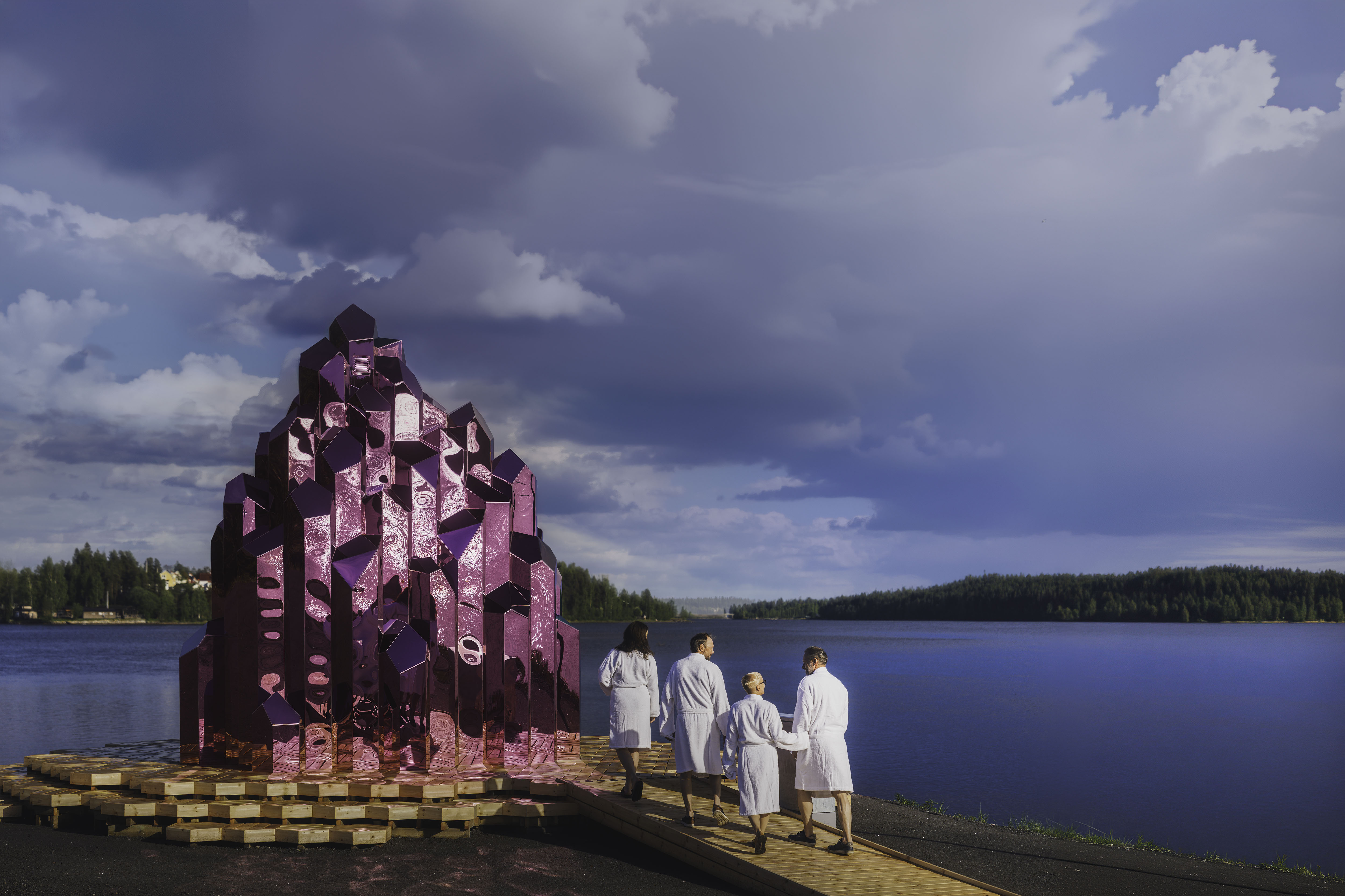 Vier mensen in badjassen lopen onder een bewolkte hemel richting de roze, kristalvormige Crystal Sauna aan de waterkant.