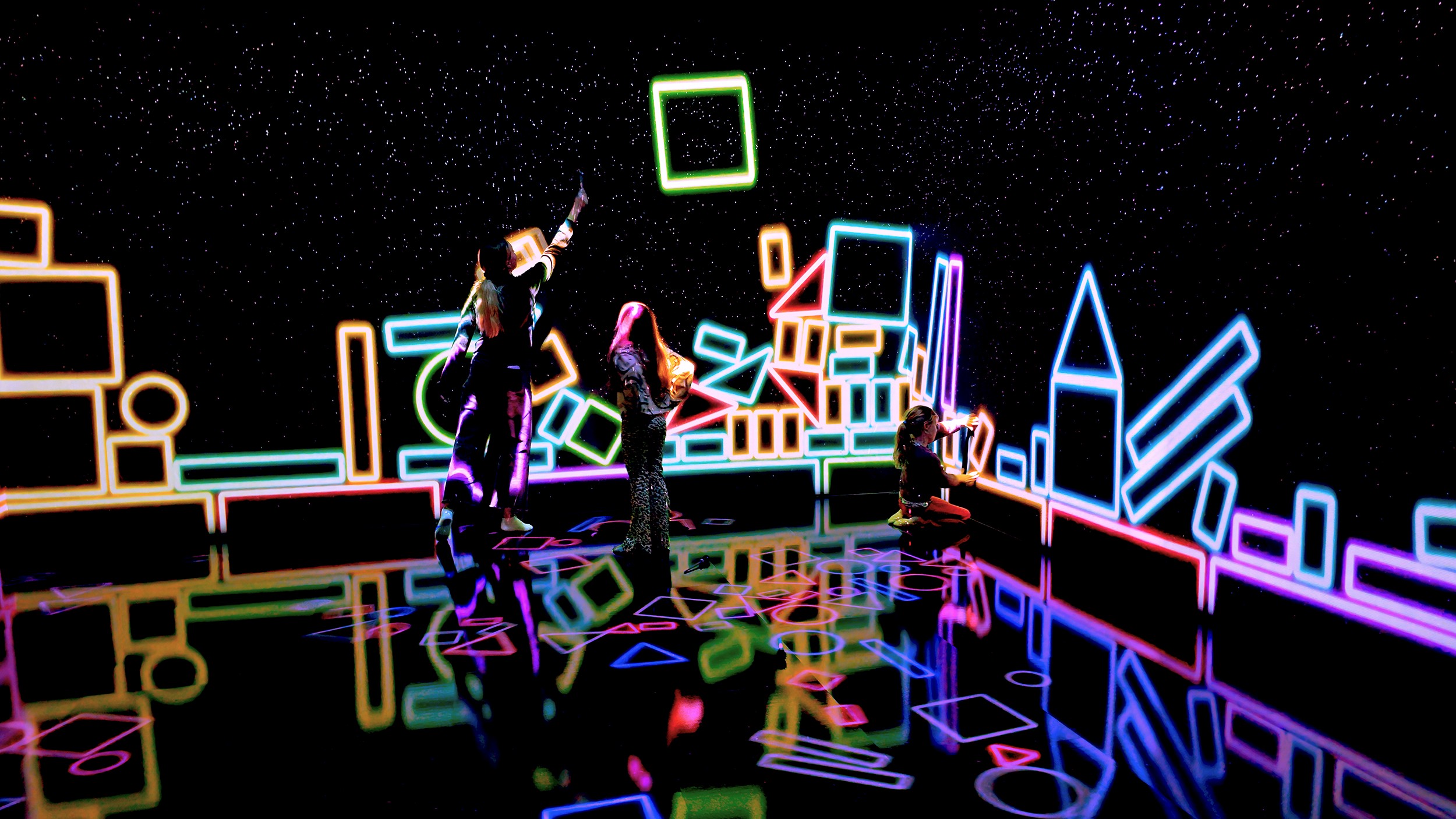 Three visitors explore an interactive digital art installation at Cube of Art, surrounded by colourful neon shapes projected onto walls and floor.