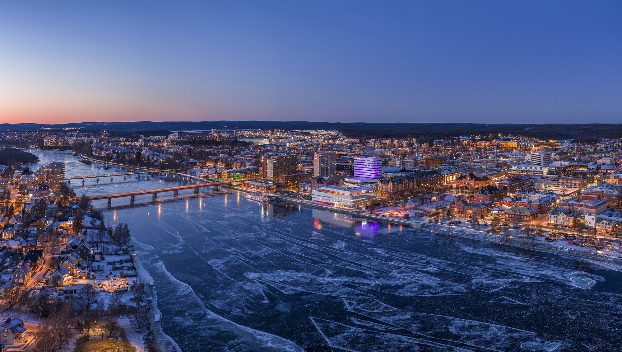 Aerial view of central Umeå in winter, with the frozen Ume River and snow-covered streets illuminated at dusk.