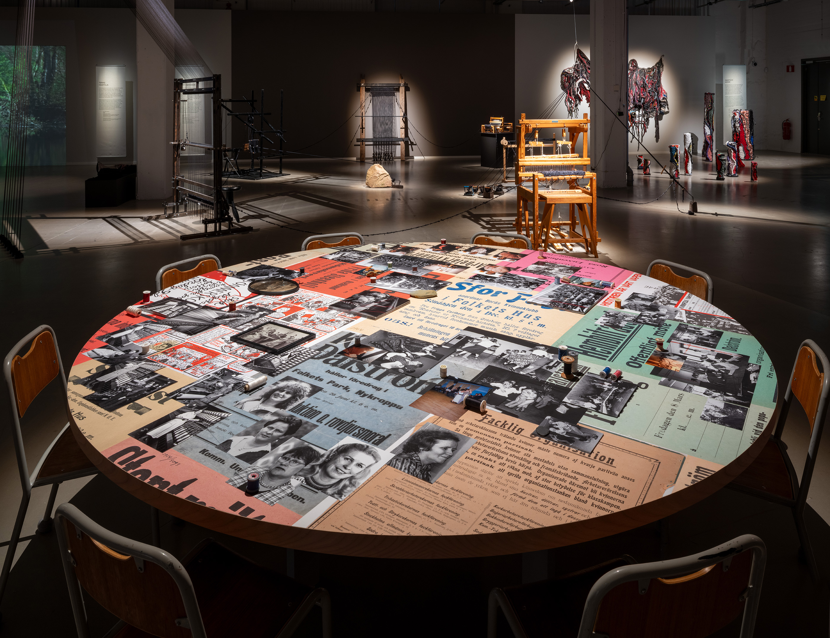 A round table covered with newspaper clippings and photographs, surrounded by chairs, with looms and colourful textile artworks behind it.