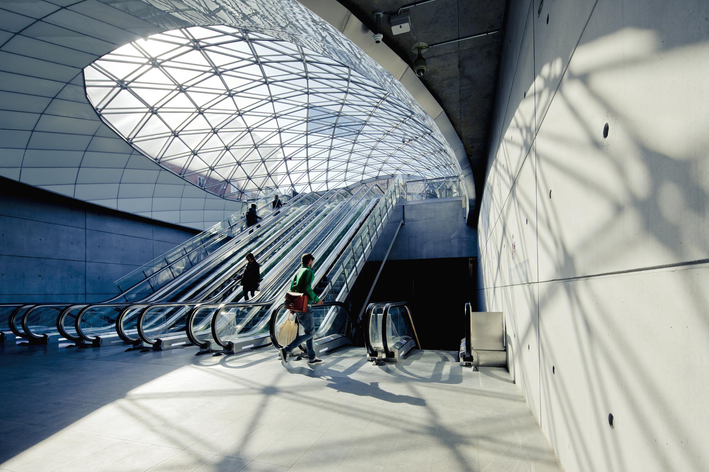 Een ondergronds station met een moderne architectuur, een grotvormige entree met een beglazingssysteem waardoor het licht de perrons binnenstroomt. Sommige mensen gaan op en neer in vier roltrappen die vanaf de ingang naar beneden leiden.