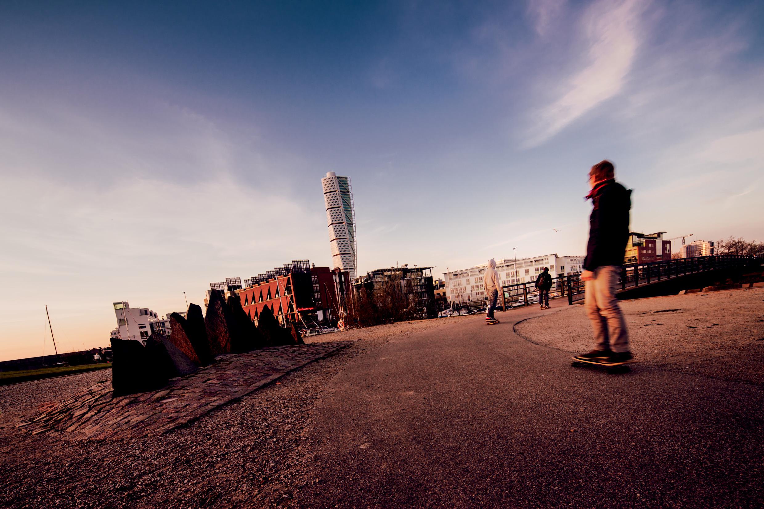 Skateboarders go down a path in Malmö with the Turning Torso skyscraper in the background.