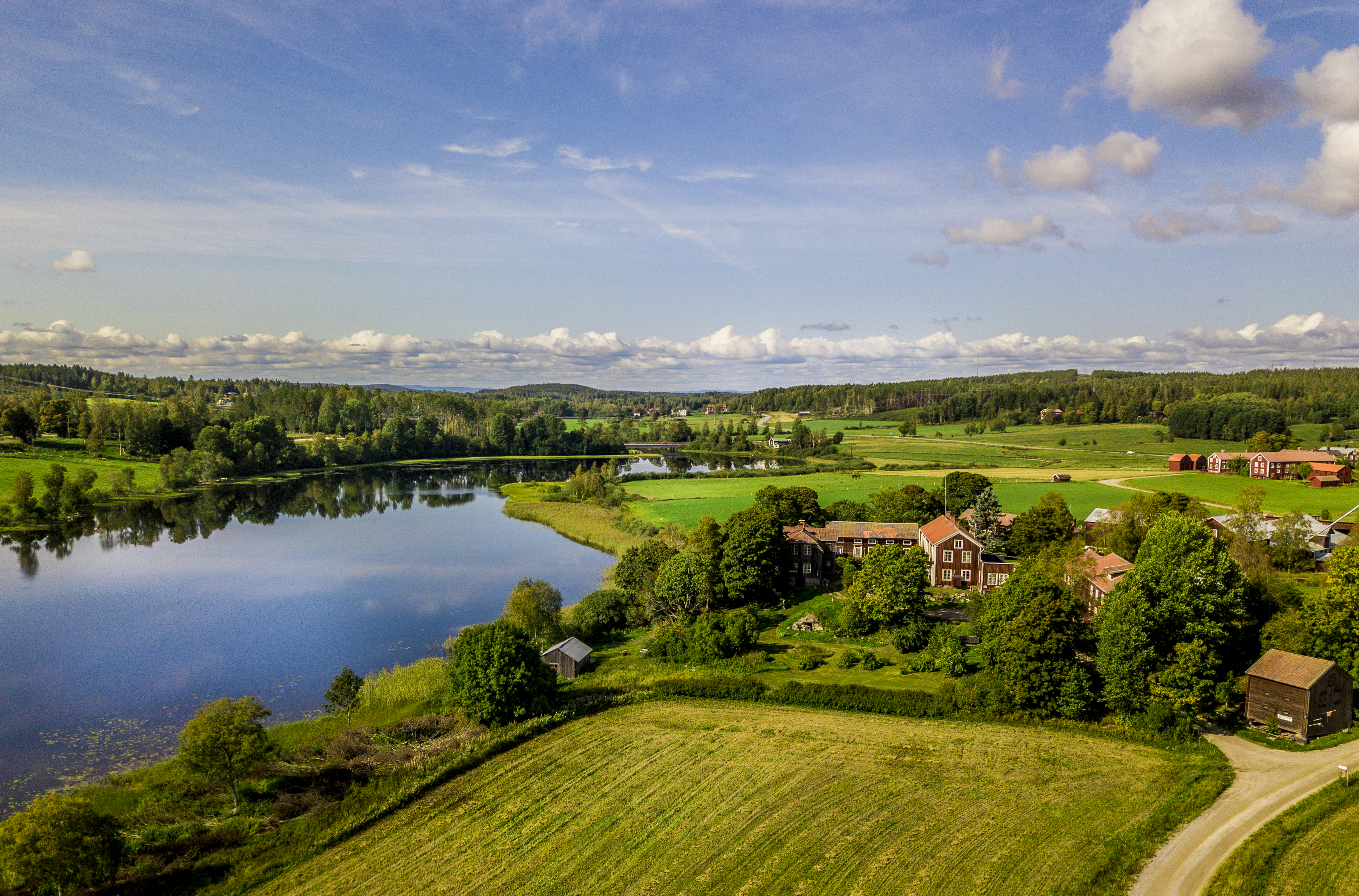 Aerial view of Frägsta Hälsingegård with red timber buildings surrounded by green fields, trees and a reflective river landscape.