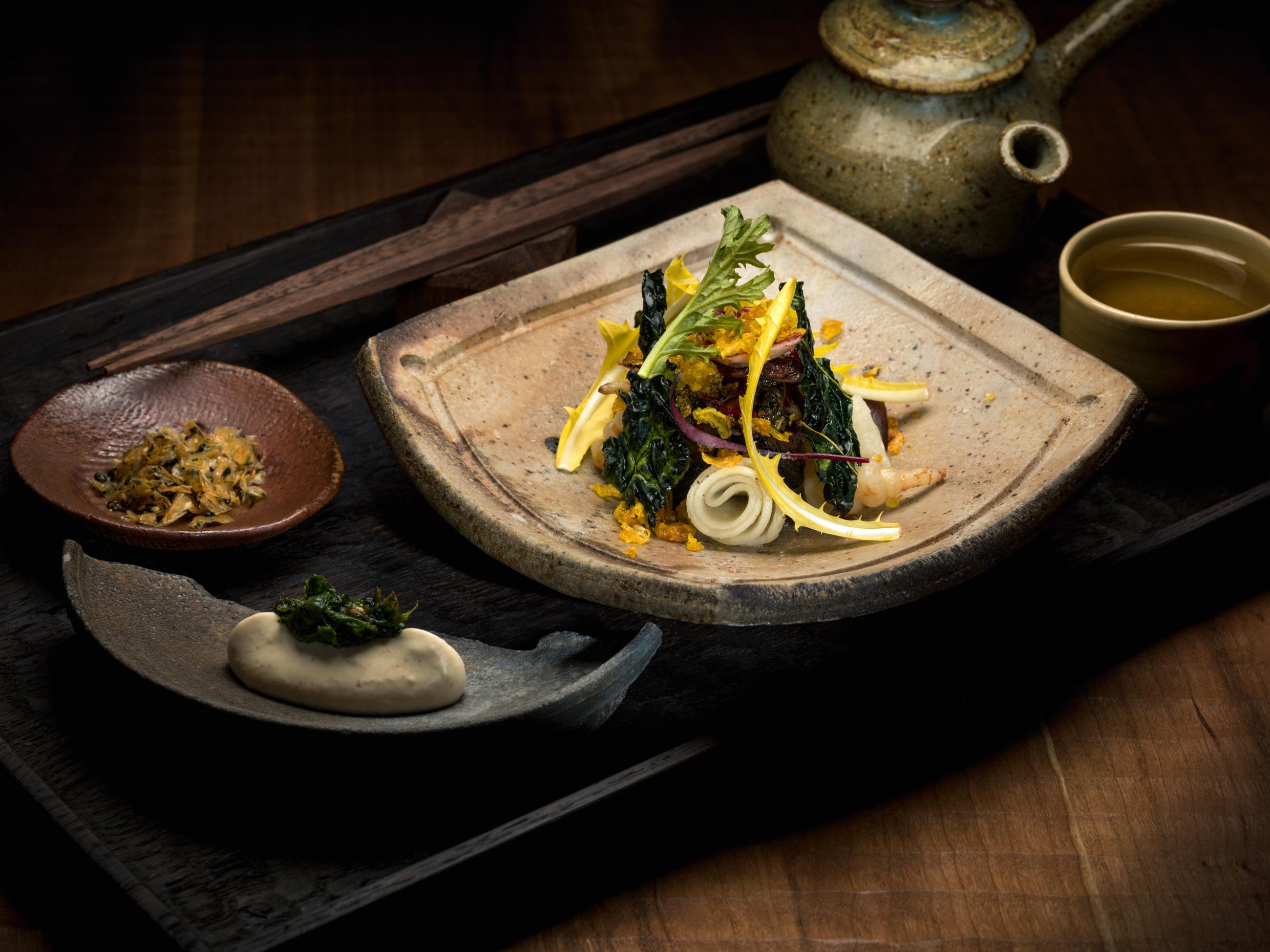 Nahaufnahme auf ein Gericht in einem gehobenen Restaurant. Ein quadratischer Teller mit einer bunten Schale in Grün und Gelb. Zwei kleine Teller mit einer Soße und einer Beilage und einer Tasse Tee.