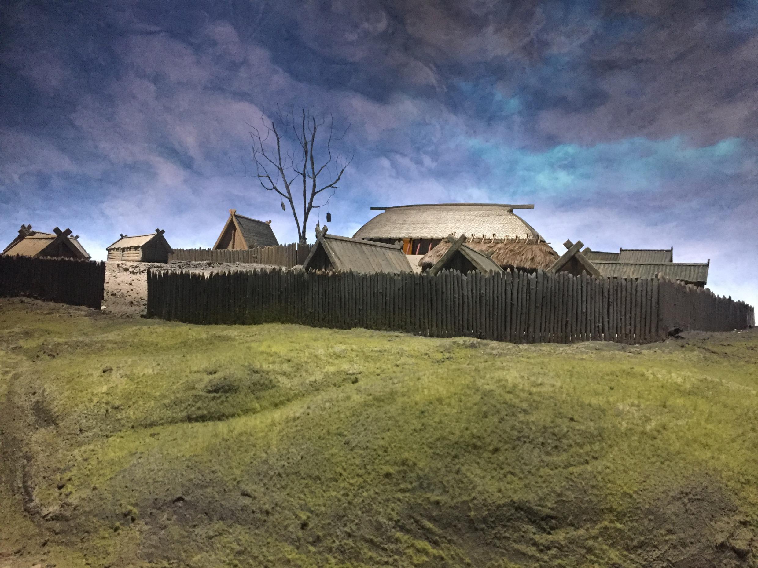 Ein Dorf aus Holzbauten hinter einem Holzzaun, davor ein bemooster Hügel