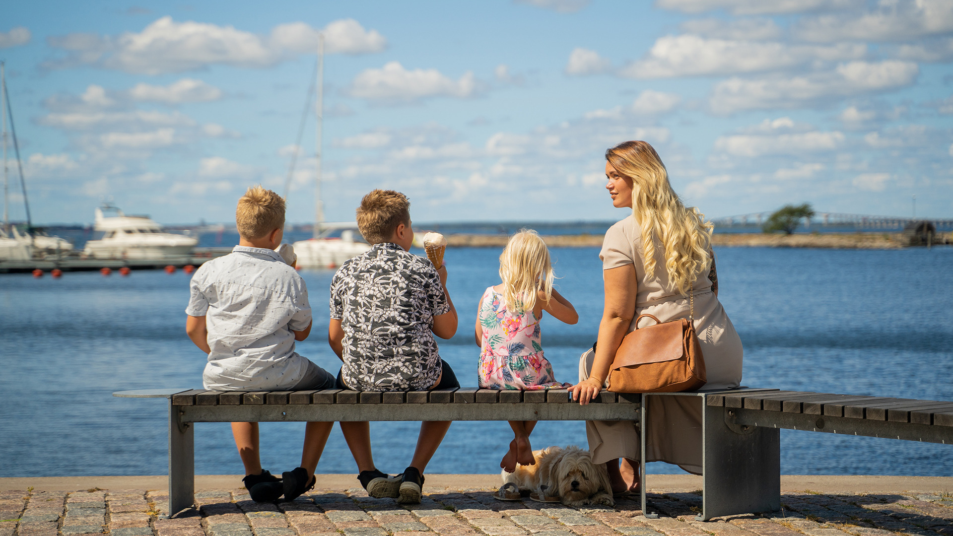 Drie kinderen en een volwassene zitten op een bankje aan zee in Färjestaden, Öland, en genieten van een ijsje met aangemeerde boten en een brug op de achtergrond.