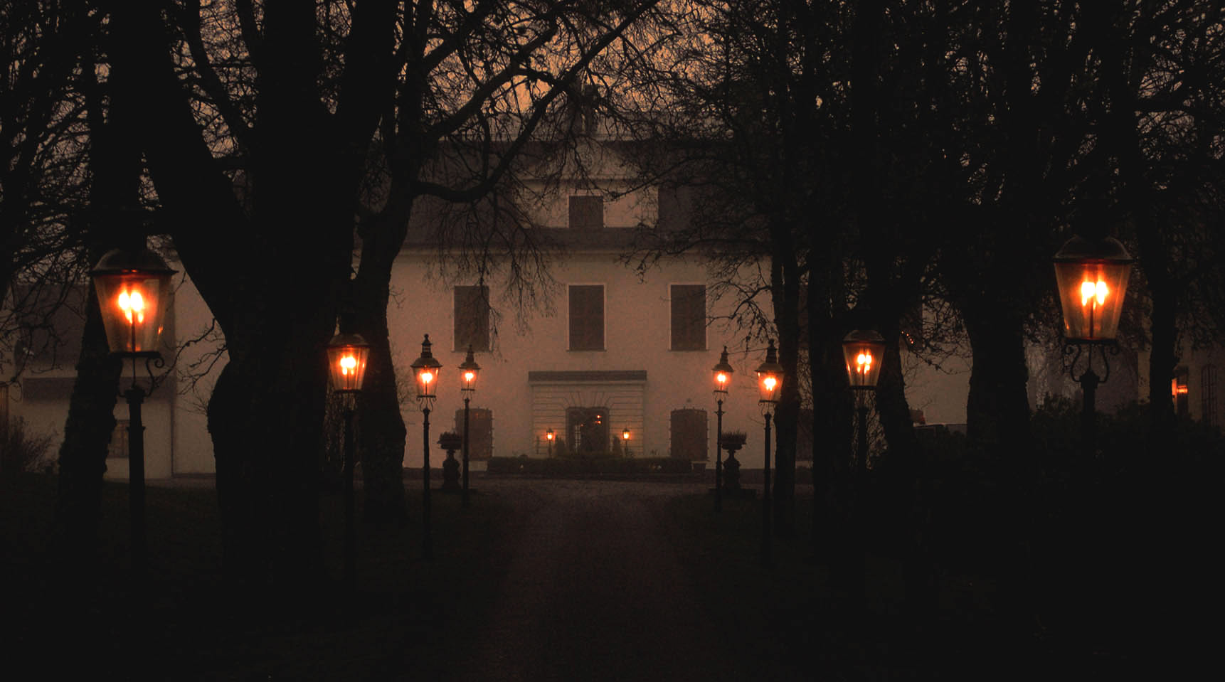 Häringe Castle in de schemering, met gloeiende lantaarns langs het pad en bomen die zich aftekenen tegen de donkere lucht.