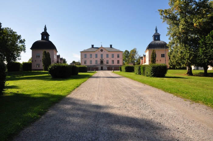 Le château de Hesselby, un manoir rose pâle, vu depuis une longue allée de gravier, avec ses deux tours rondes symétriques, ses pelouses verdoyantes et ses arbres, par une journée ensoleillée.