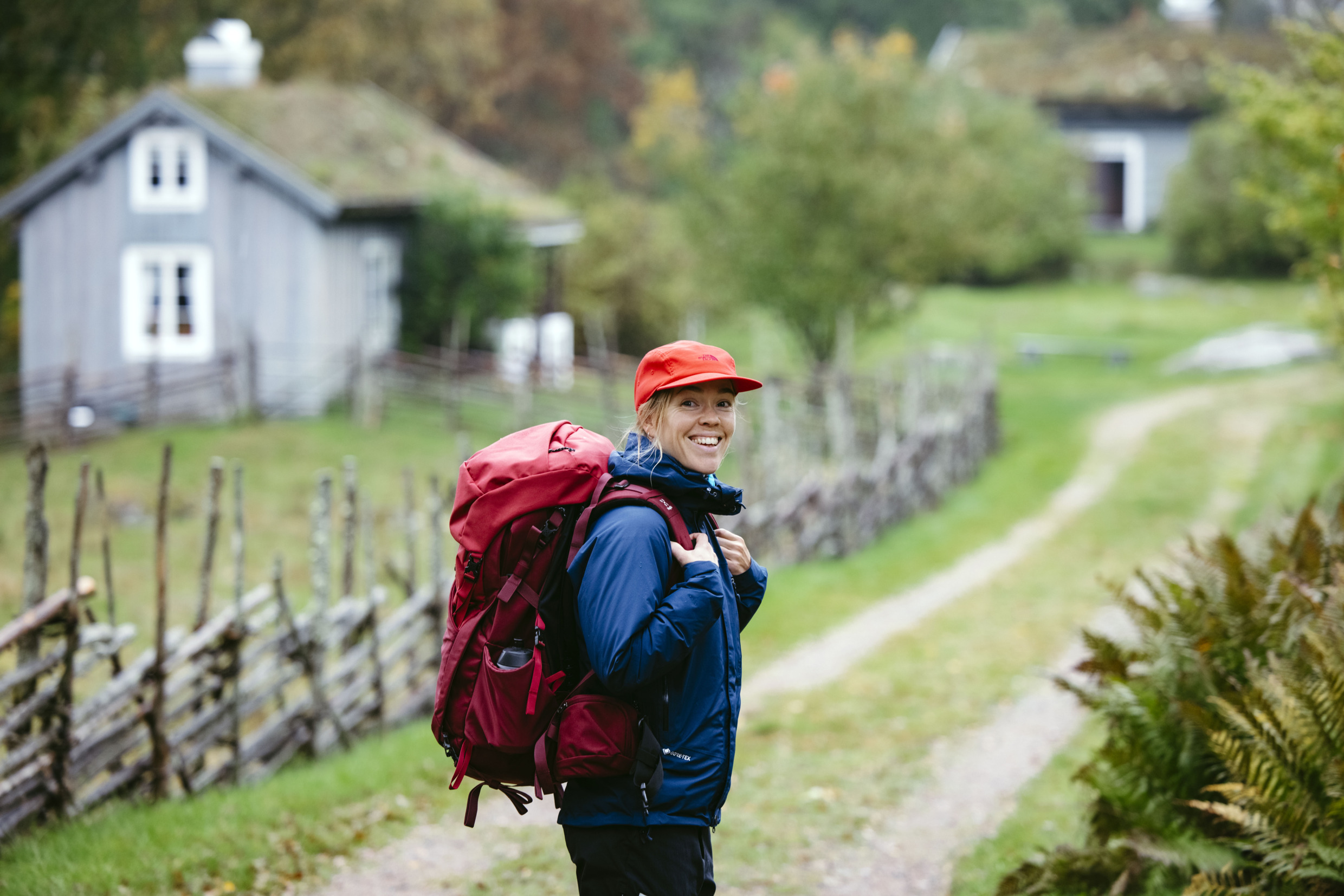 Eine Frau in Outdoor-Kleidung und mit Rucksack schaut in die Kamera, während sie auf einem Wanderweg steht. Im Hintergrund ist ein Holzhäuschen zu sehen.
