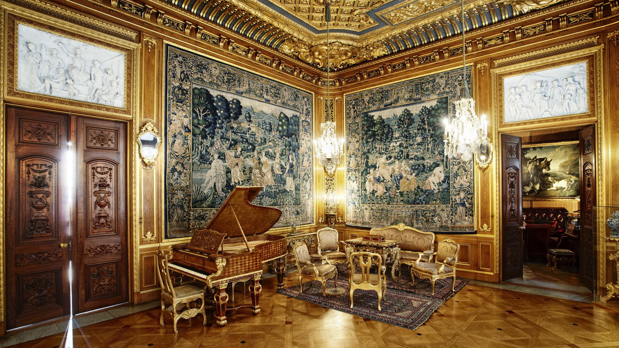 The luxurious, golden sitting room of Hallwyl house, with large paintings, a grand piano, golden furniture and chandeliers.