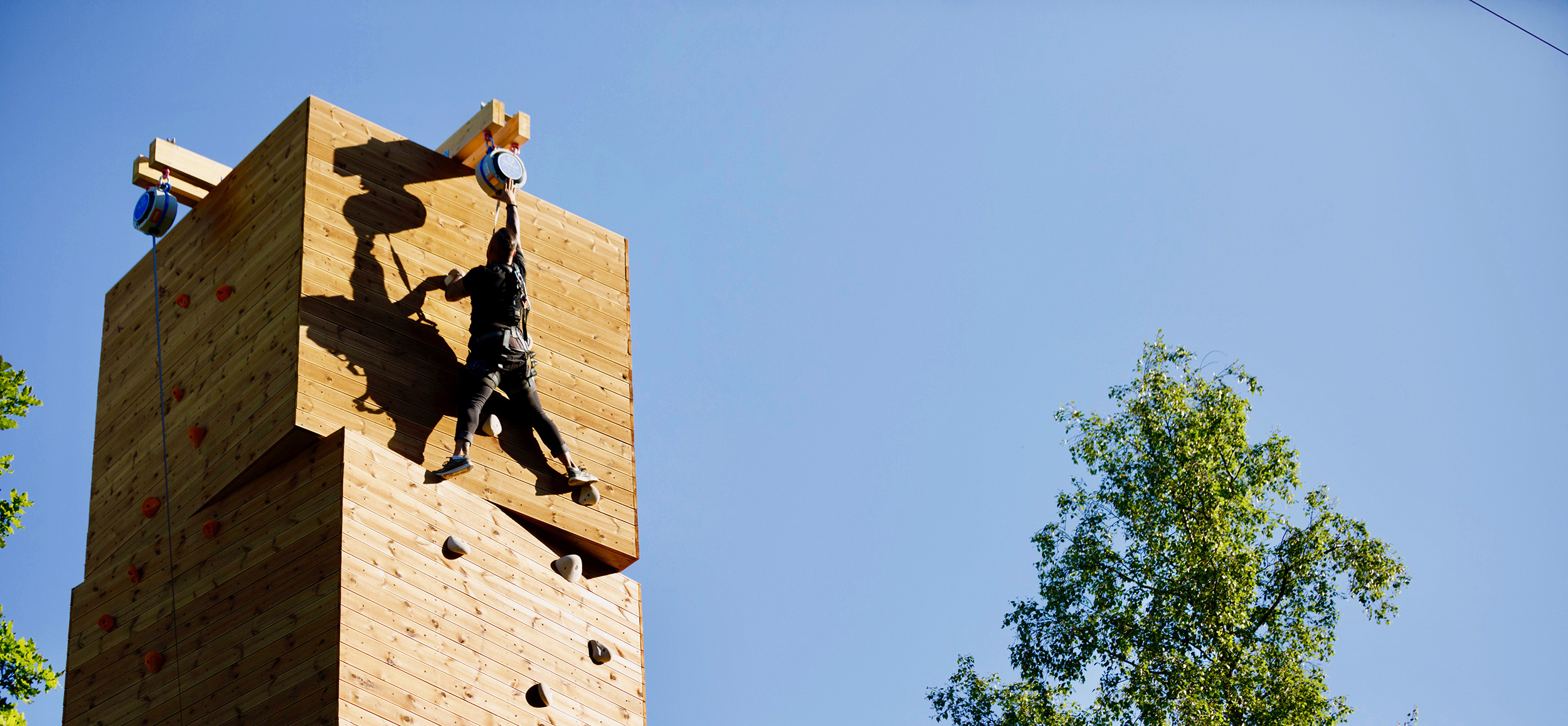 A person climbing a tall wooden climbing tower at Kungsbygget Adventure Park, reaching for the top against a clear blue sky.