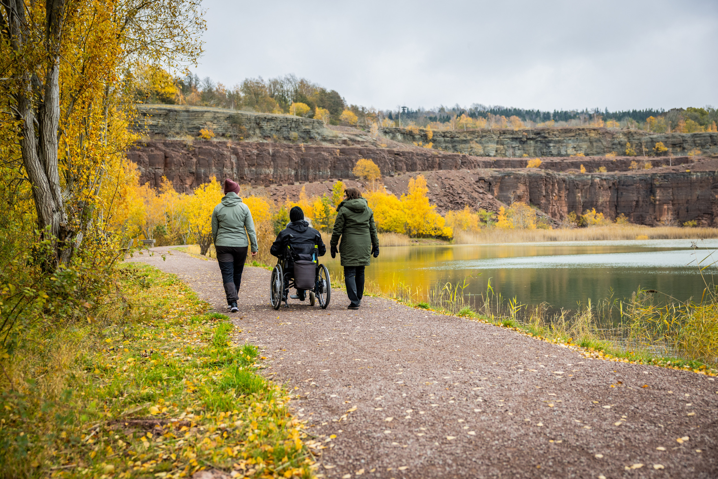 Three people, one of whom is in a wheelchair, walk by the Kinnekulle quarry on an autumn day.