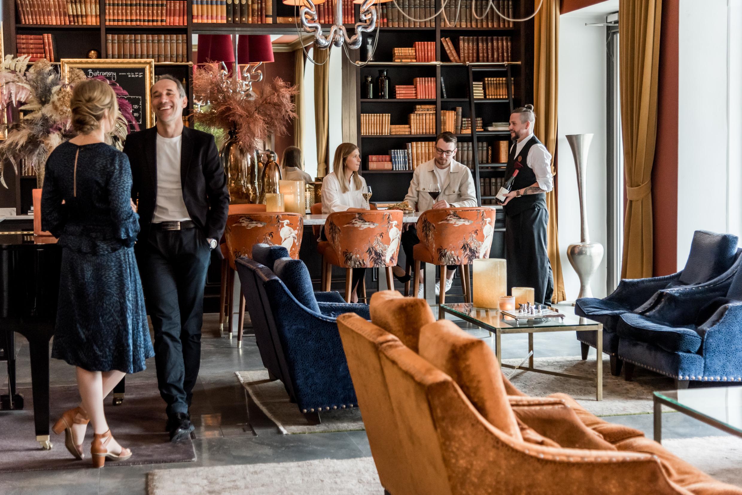 Des personnes dans un lobby d'hôtel. Le lobby est décoré avec des livres, des canapés en cuir de différentes couleurs, des tables et des vases.