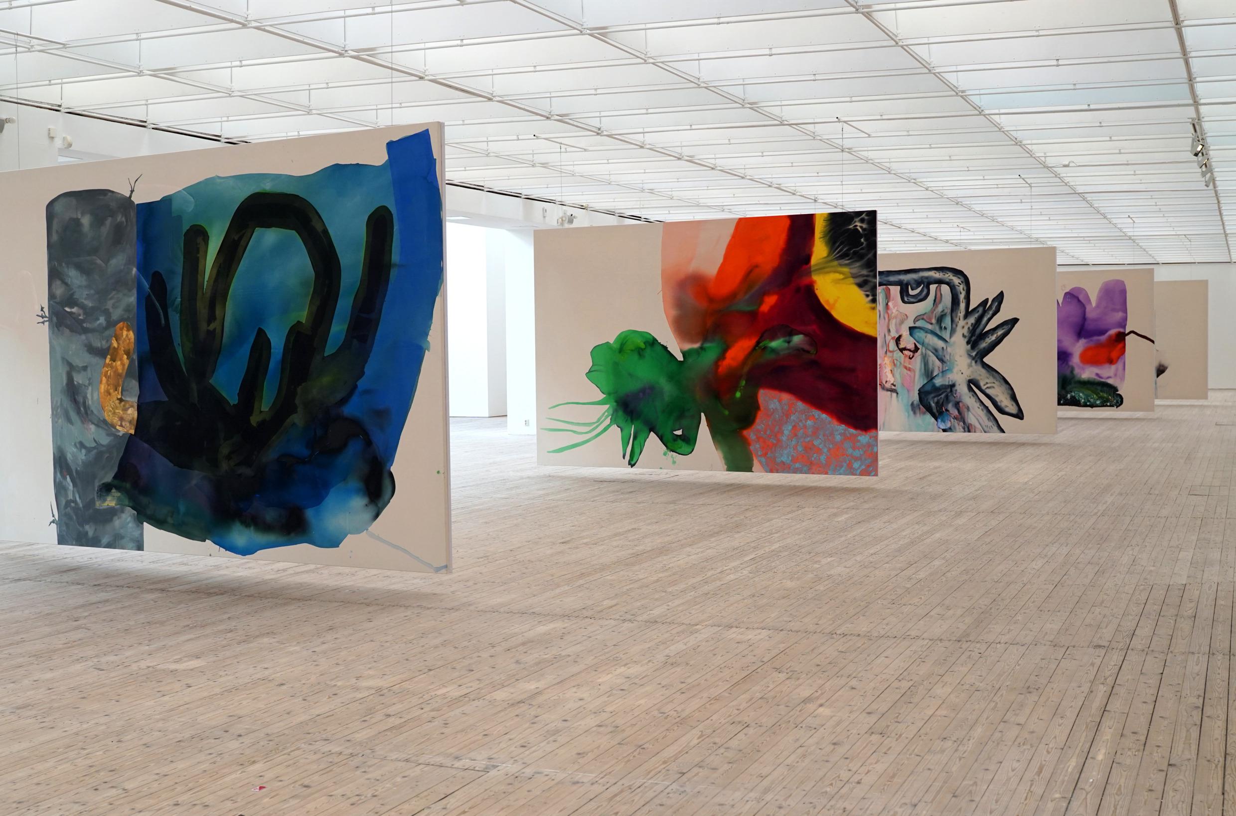 Several large paintings are hanging from the ceiling in the middle of a large exhibition hall.