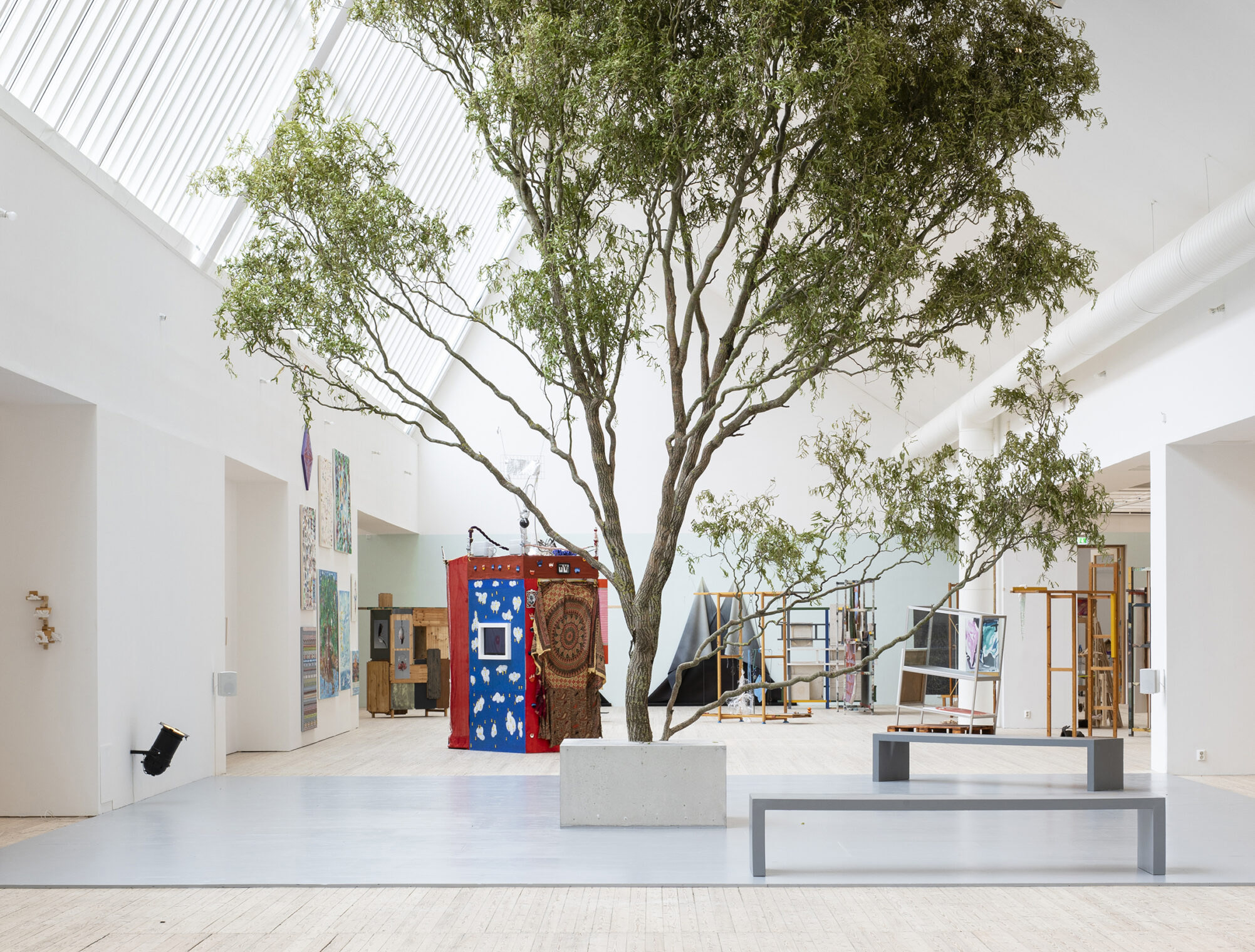 A large tree grows inside Malmö Konsthall’s bright exhibition space, surrounded by colourful artworks and benches beneath a skylit ceiling.