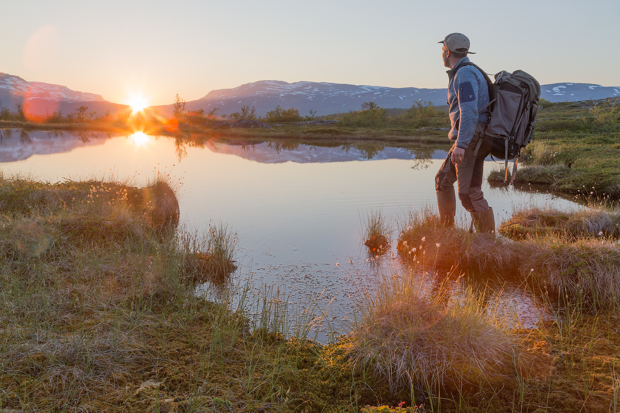 Een man met een rugzak staat naast een meer terwijl hij naar de middernachtzon kijkt.