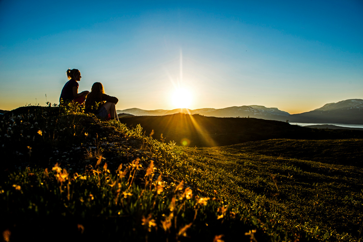 Twee mensen zitten op een met gras begroeide heuvel en kijken naar de middernachtzon boven de bergen en een meer in Noord-Zweden.