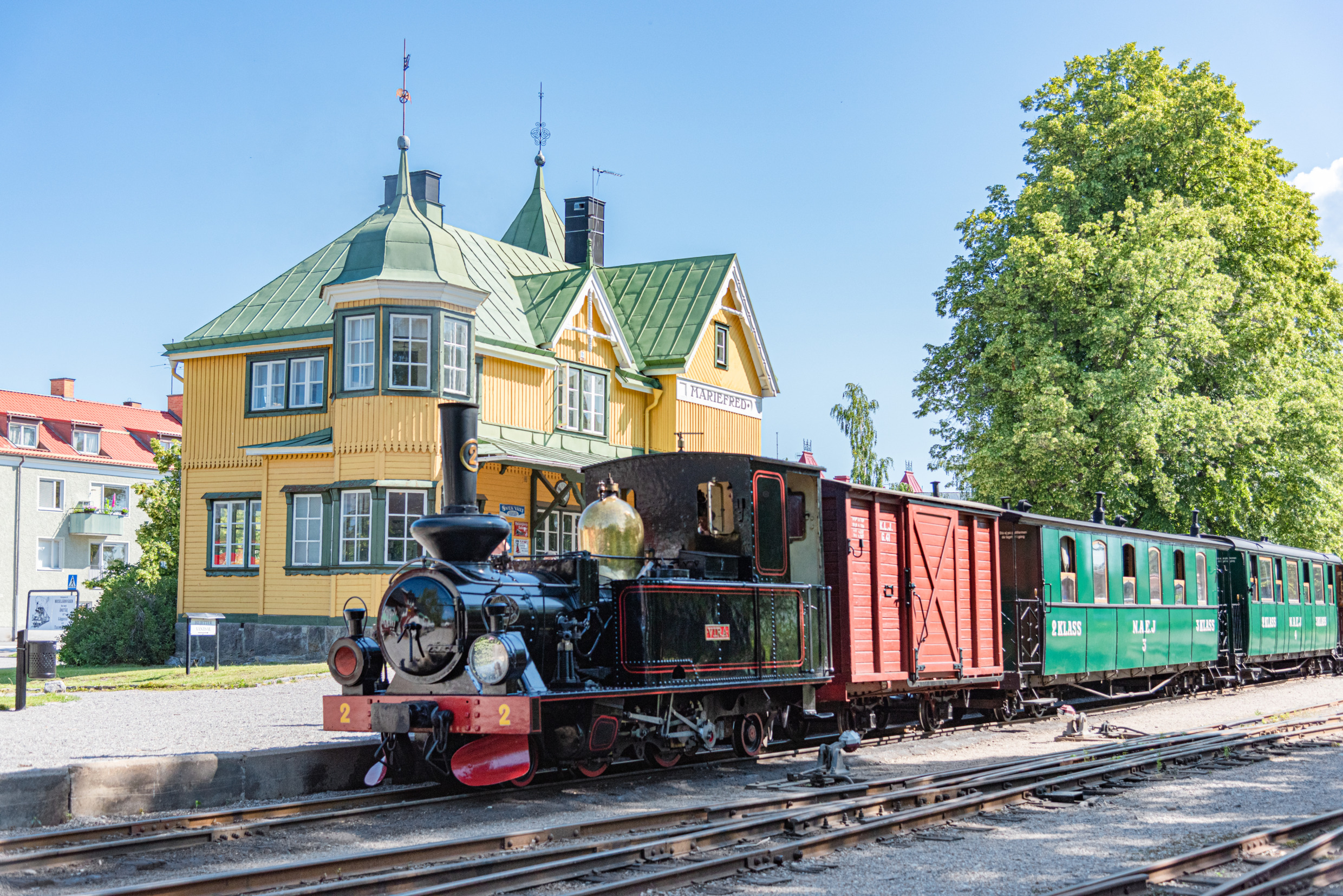 Historischer Dampfzug mit dem alten Bahnhofsgebäude in Mariefred im Hintergrund.
