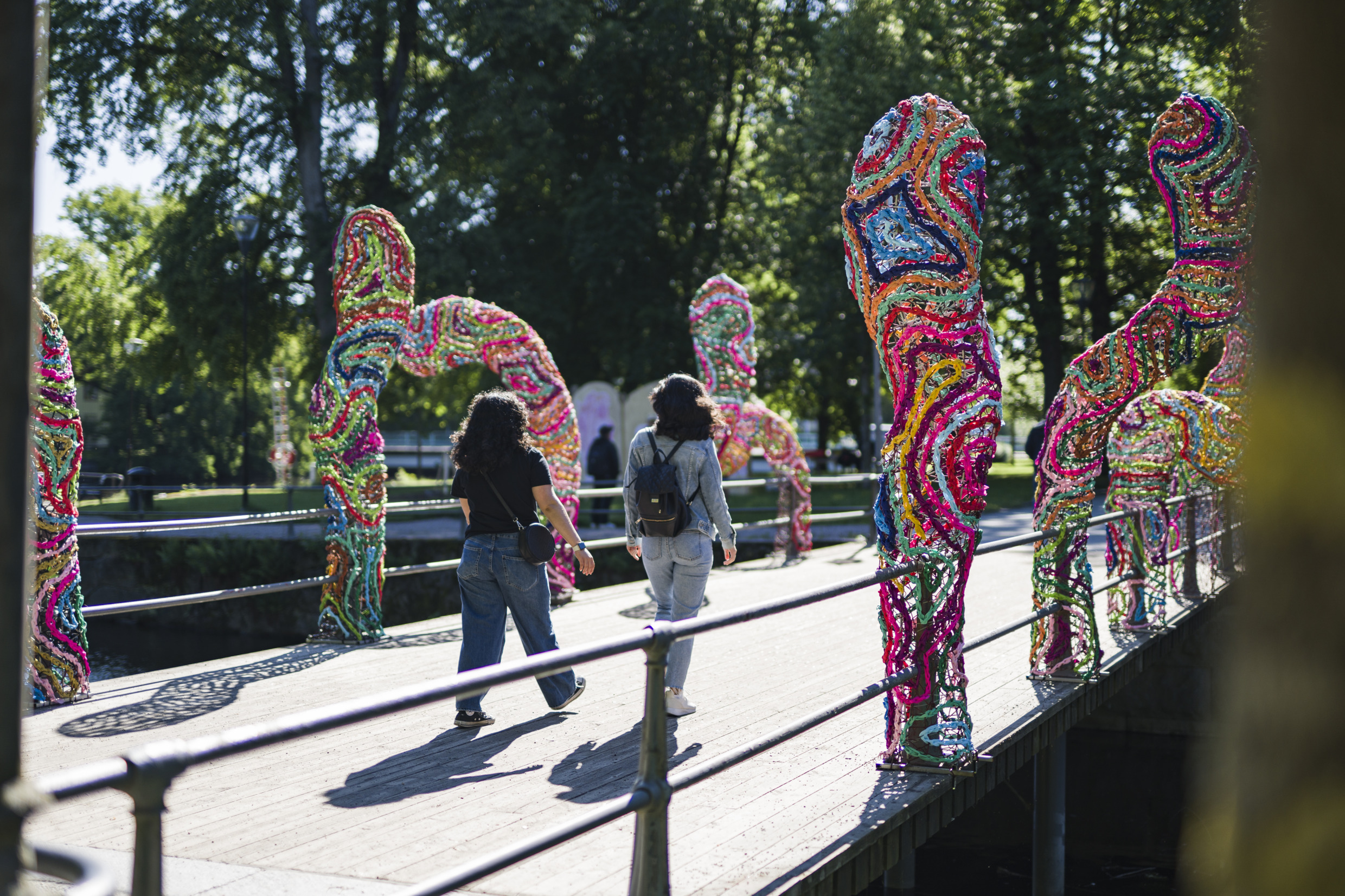 Twee vrouwen lopen over een brug met een kleurrijke kunstinstallatie aan de zijkanten.