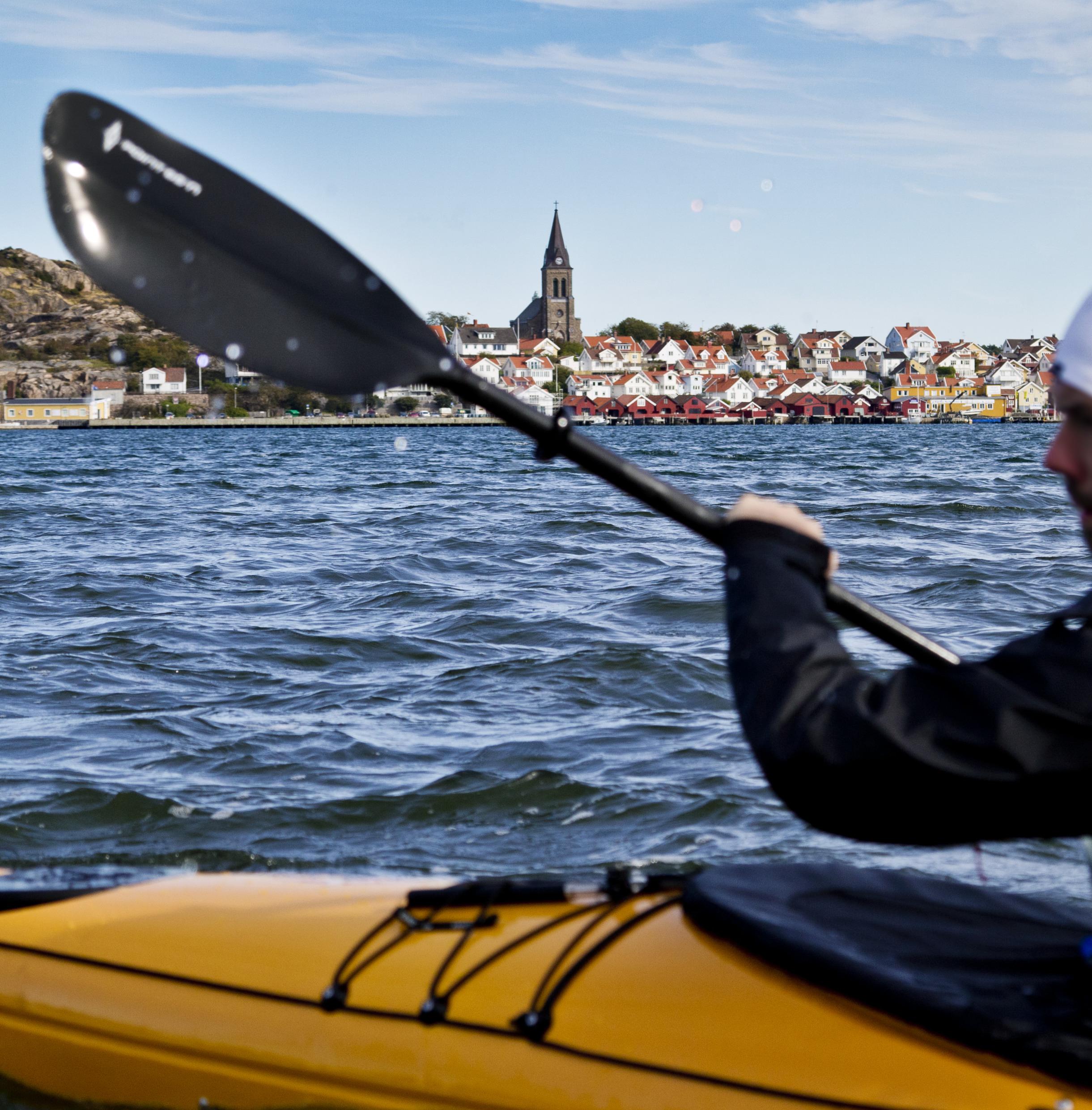 Im Vordergrund paddelt ein Mann in einem gelben Kajak, im Hintergrund der Küstenort Fjällbacka mit seinem markanten Kirchturm.