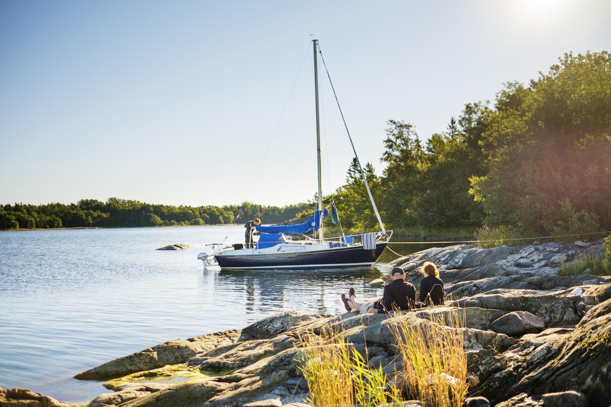 Een zeilboot met een man erop ligt aangemeerd aan een klif in de archipel. Een man en een vrouw zitten op de kliffen.