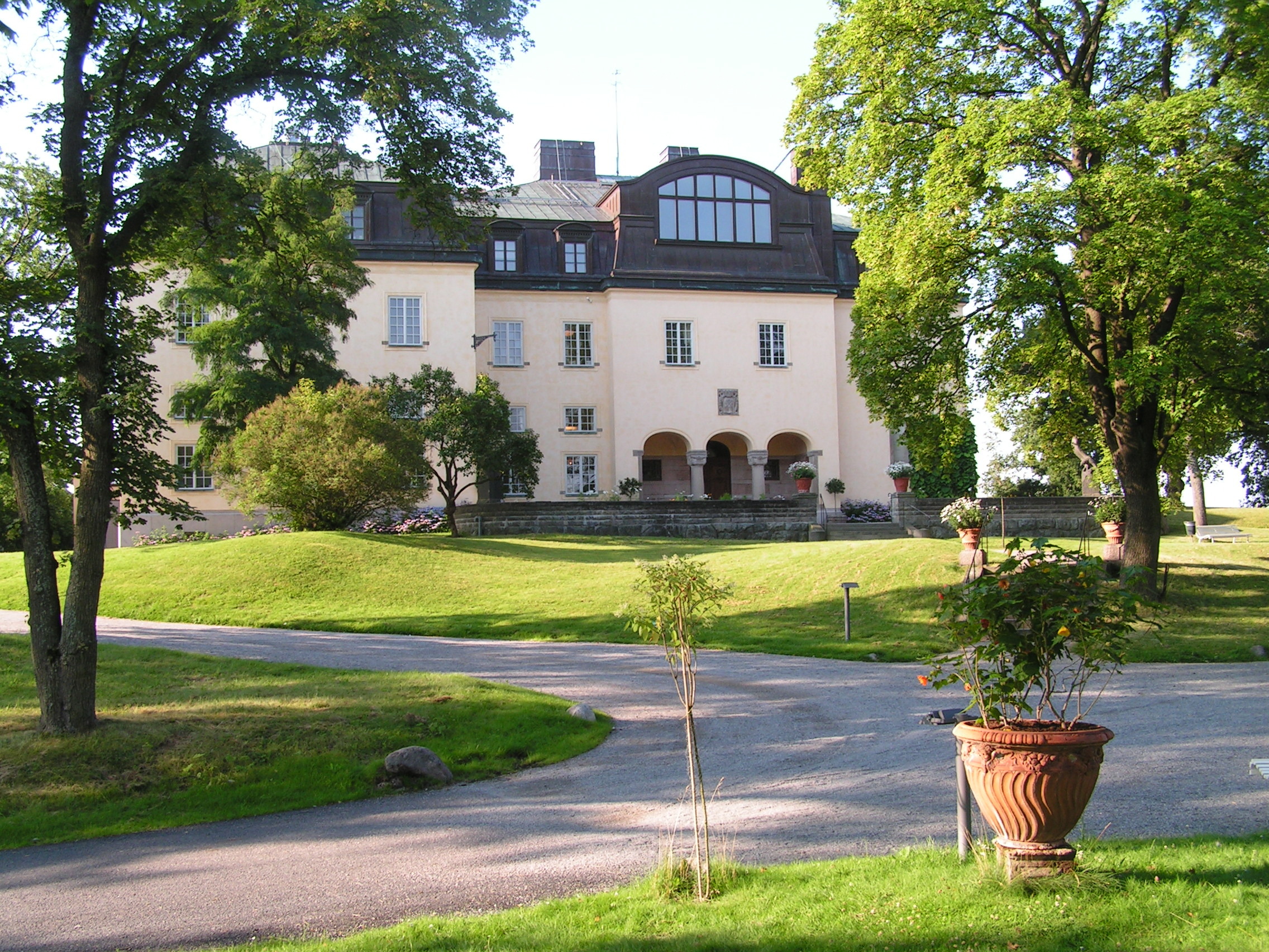 The façade of Prince Eugen’s Waldemarsudde in Stockholm, a light-coloured palace surrounded by lush gardens and winding paths, captured on a sunny day.
