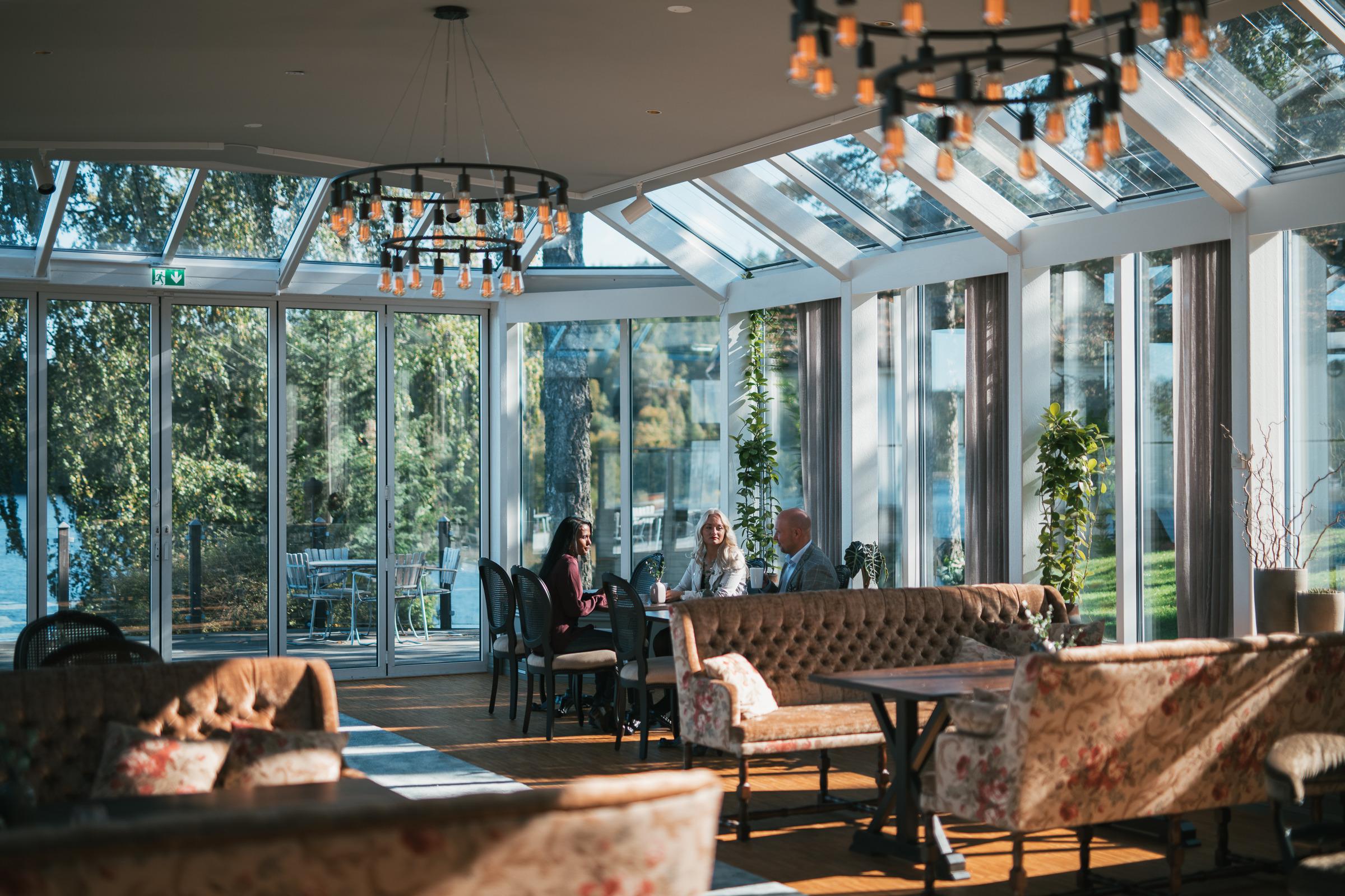 Drie personen zitten in een loungeruimte in een oranjerie met grote ramen. De kamer is ingericht met banken, stoelen en tafels.