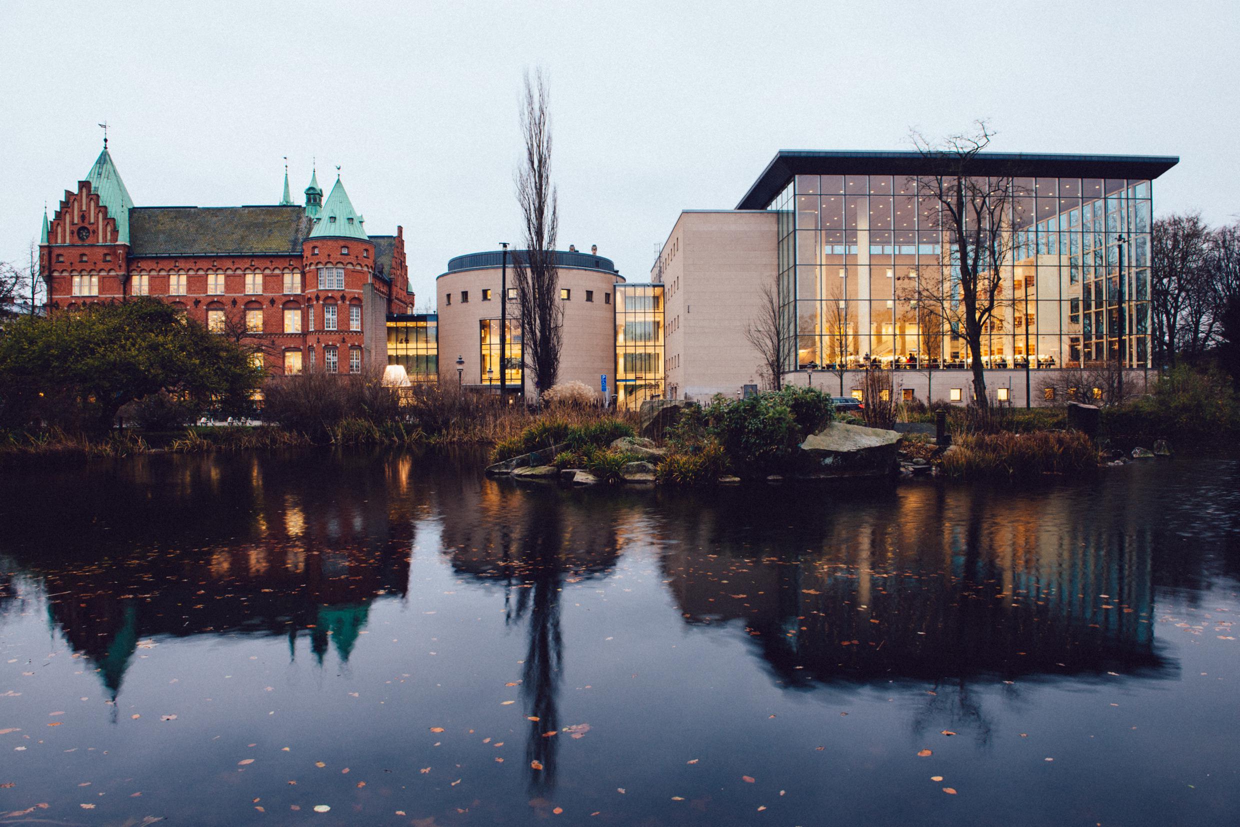 Ein altes rotes Backsteingebäude mit Türmen neben einem modernen Gebäude mit Glasfassade an einem Fluss.