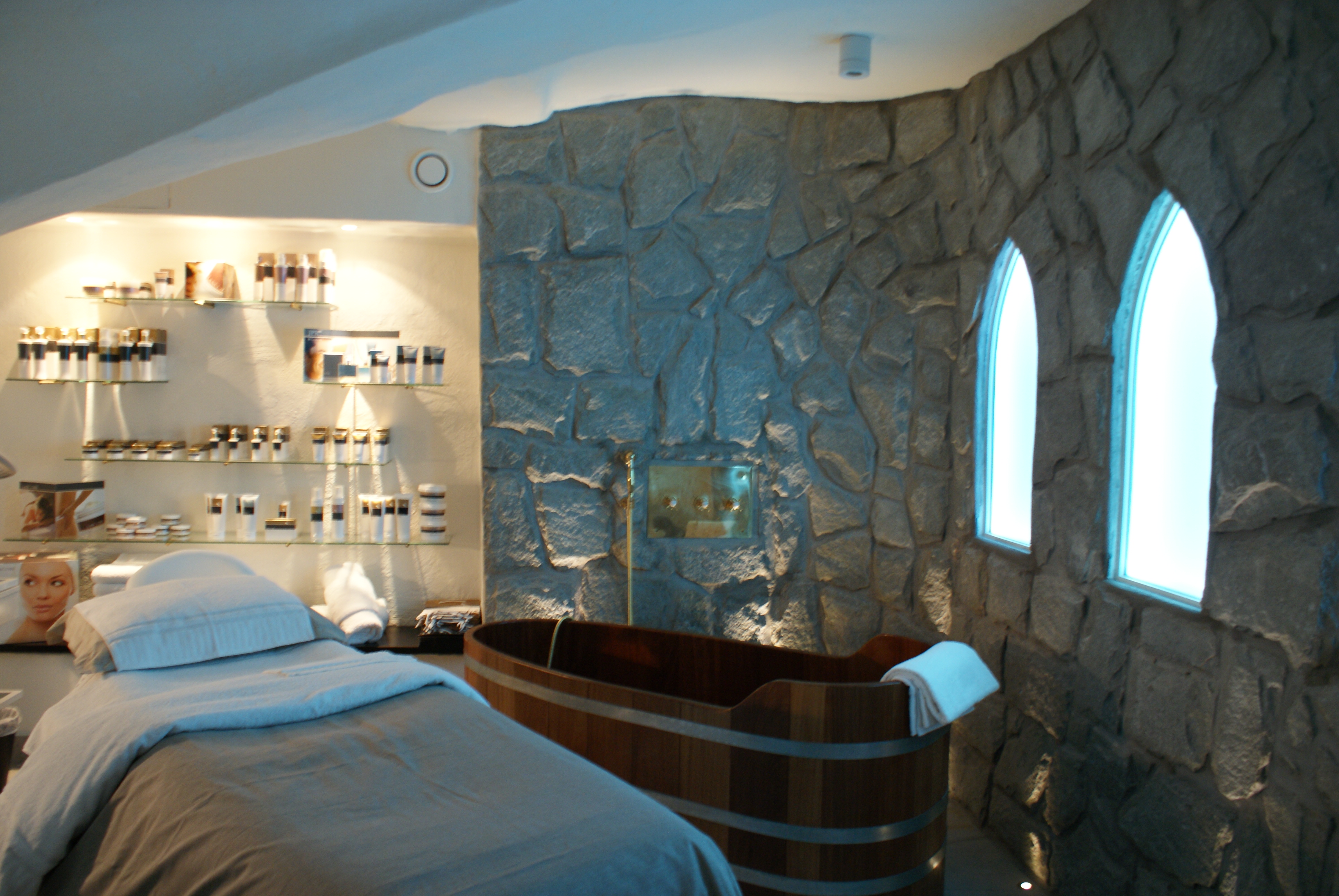 Une salle de soins apaisante, équipée d'une table de massage, d'une baignoire en bois, de murs en pierre et d'étagères faiblement éclairées sur lesquelles sont disposés des produits de soin pour la peau.