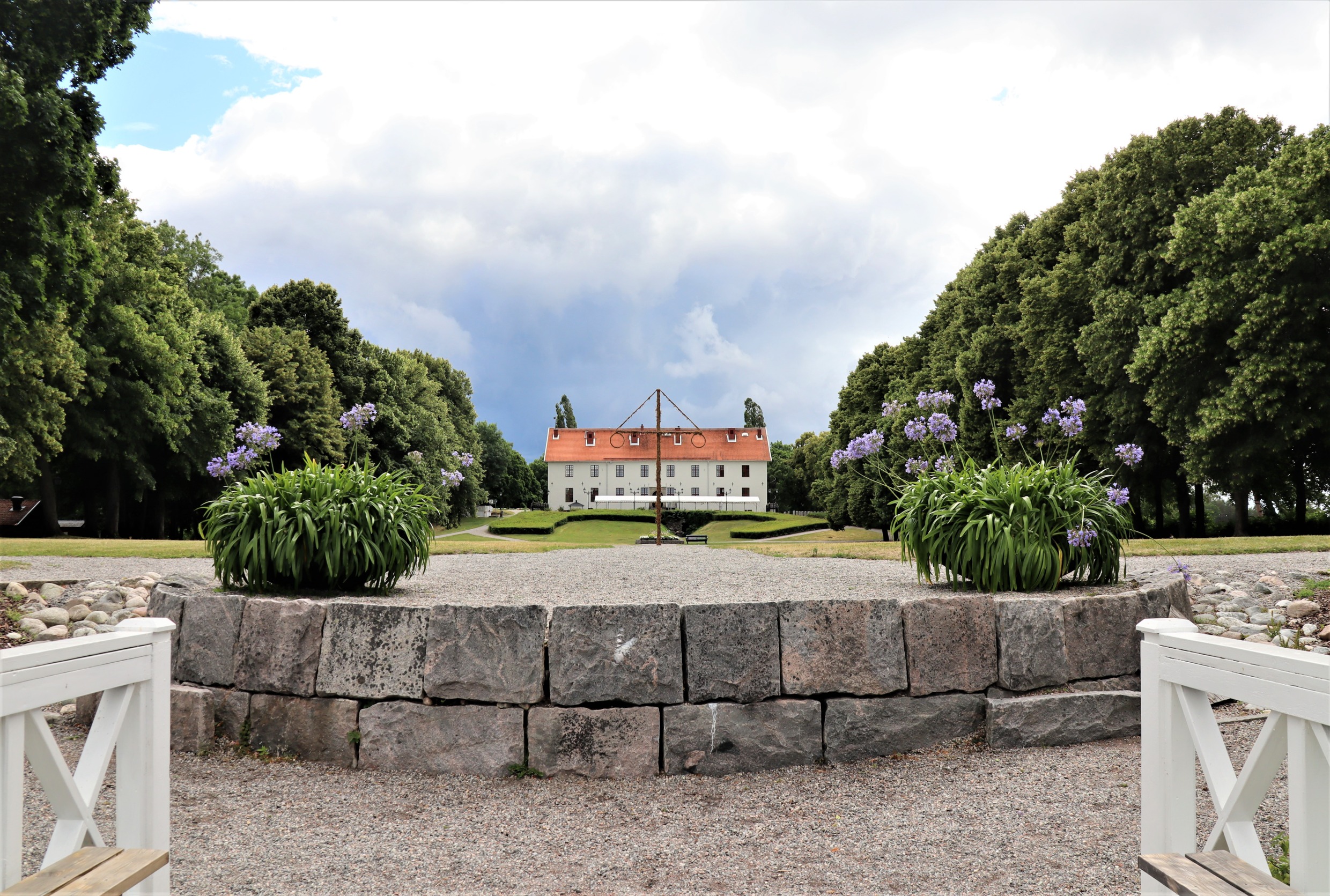 Un portail en bois blanc menant à un mur de pierre plus bas, avec le château de Sundbyholm en arrière-plan, entouré d'arbres et de buissons verdoyants.