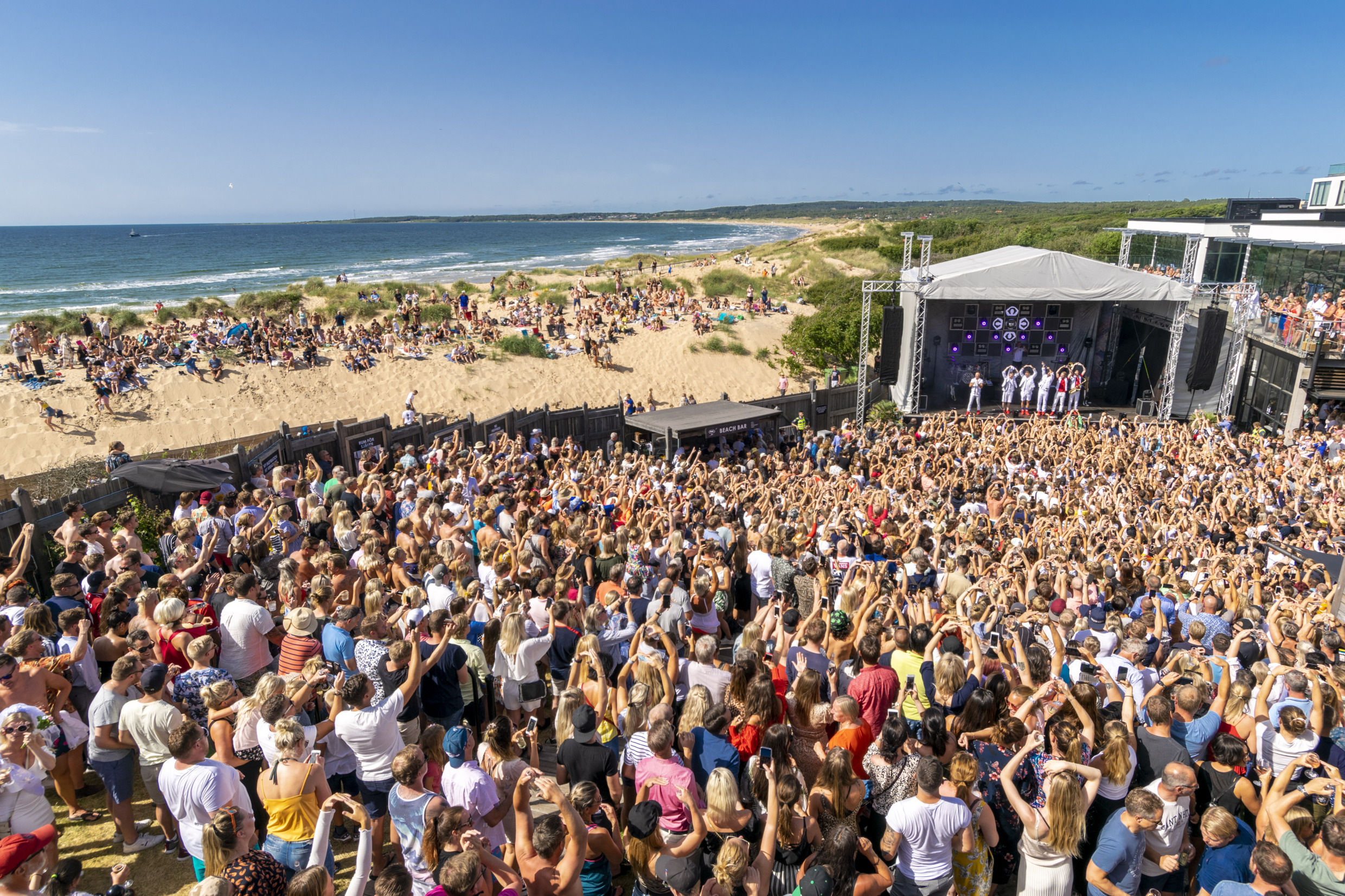 Une grande foule se tient devant une scène sur la plage à côté de la mer.