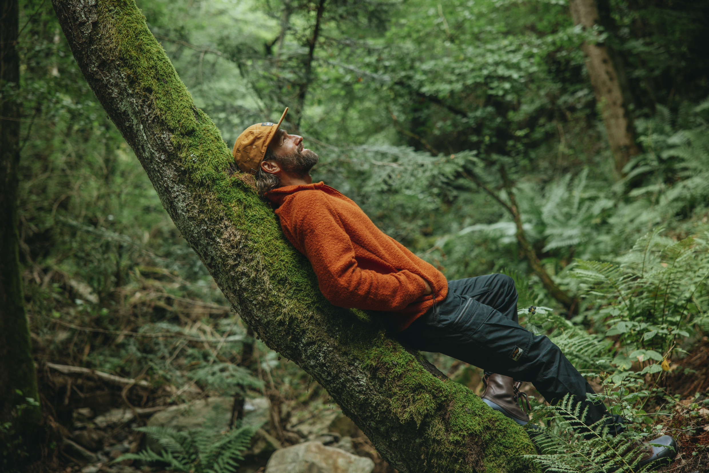 Un homme avec une veste orange et une casquette est adossé à un arbre dans une forêt.