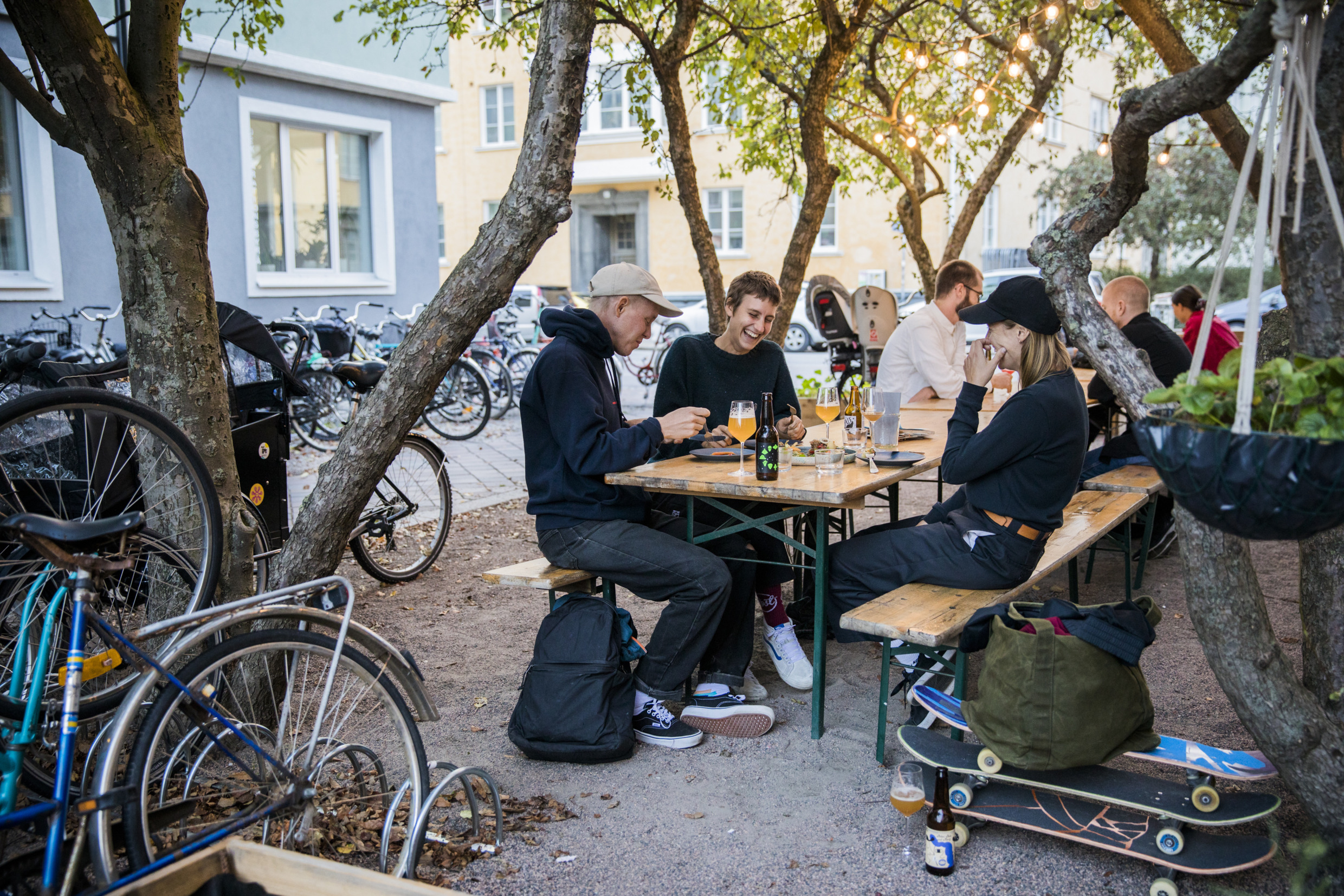 Three persons sit at a table at an outdoor restaurant laughing and eating. They have their skateboards beside the table and there are several bikes parked beside the restaurant.