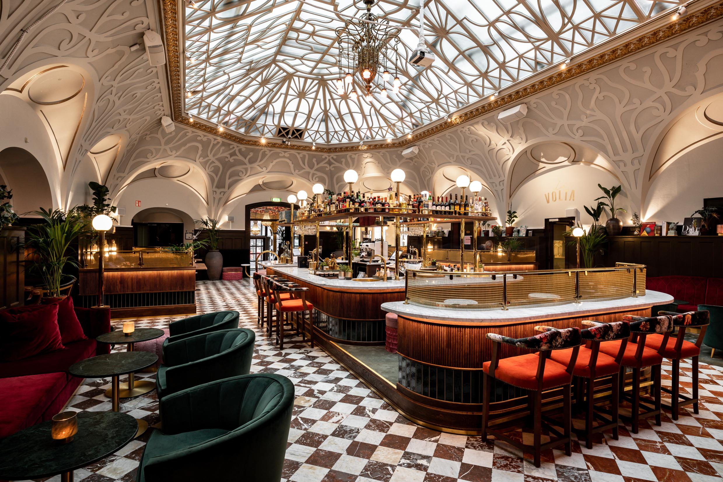 Een grote bar met een marmeren blad en een gouden standaard met allerlei flessen drank in de lobby van een hotel. Er staan barkrukken met fluwelen zittingen en links is een zithoek met een rode fluwelen bank en groene fauteuils.