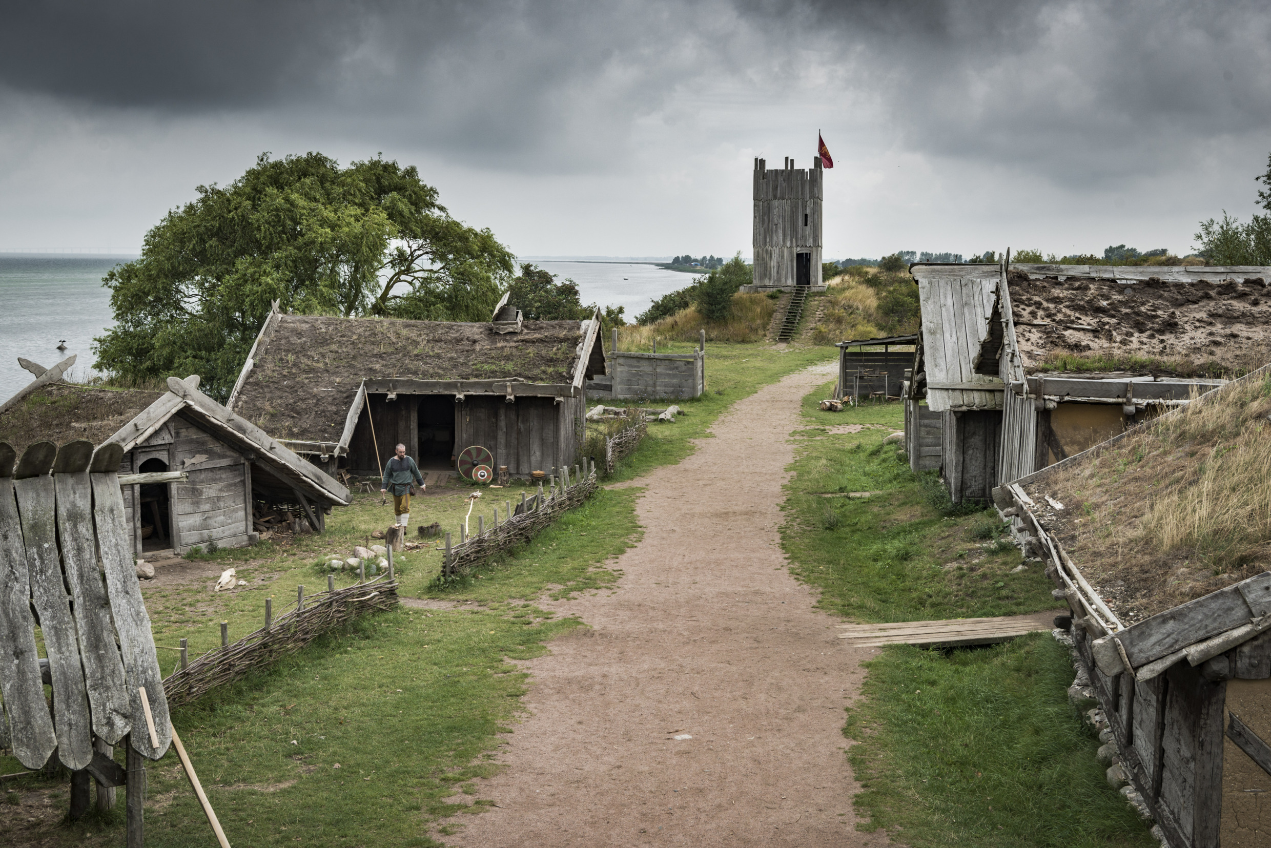 Eine Schotterstraße führt durch ein traditionelles Wikingerdorf mit Holzhäusern und grasbedeckten Dächern.