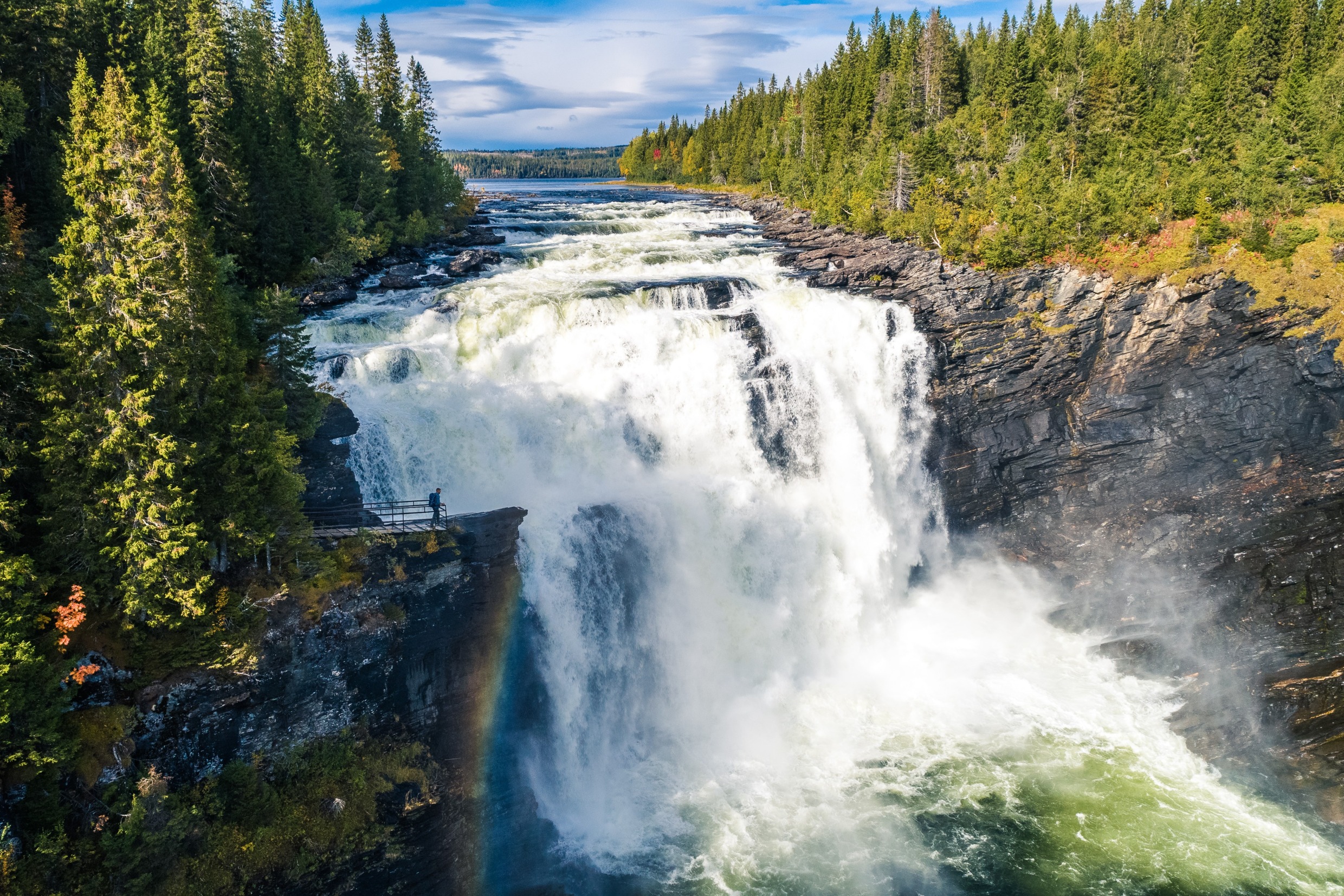 Een brede waterval omringd door sparrenbossen. Een persoon staat op een uitkijkrots aan de linkerkant van de waterval.